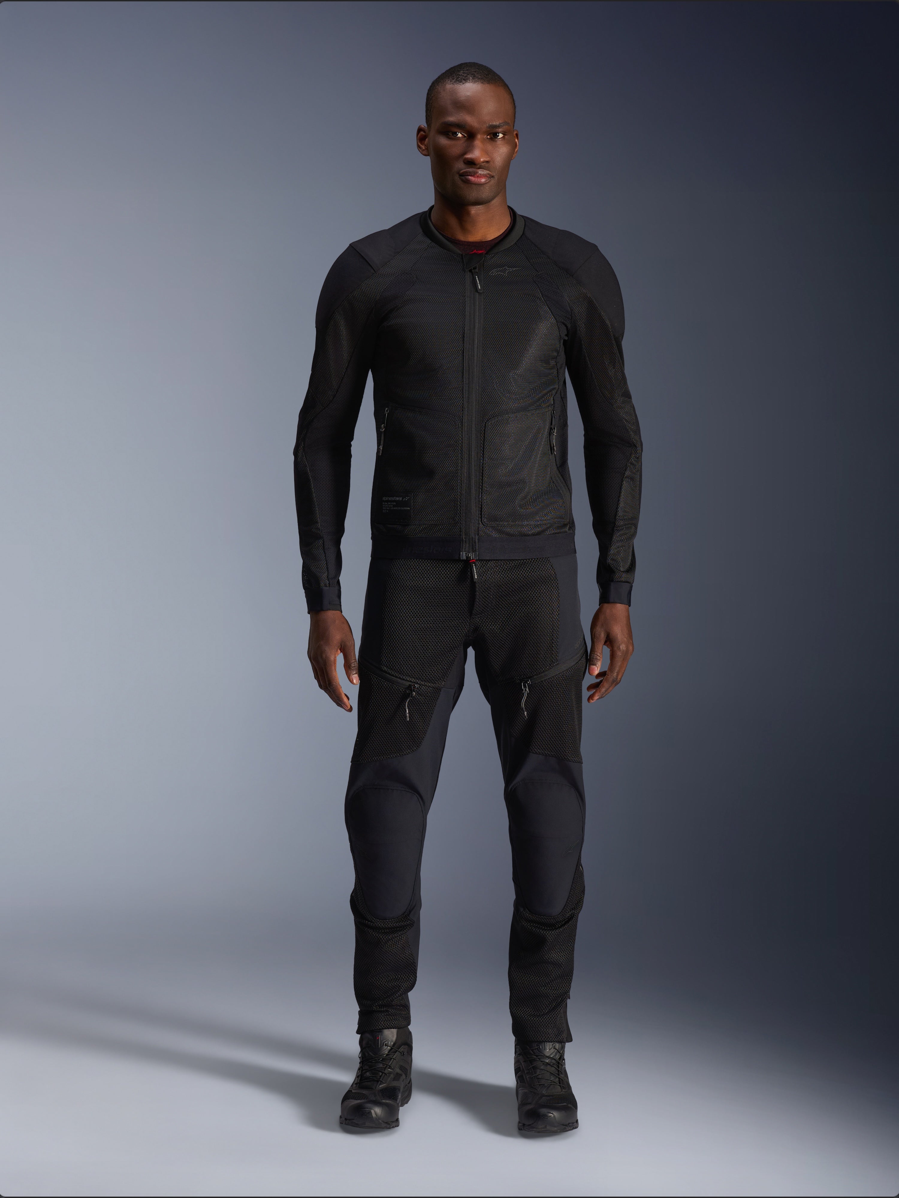 Männliches Model in der Alpinestars Troop-Air Jacke in Black Black, leichte Mesh-Motorradjacke, kombiniert mit passender schwarzer Motorradhose und Stiefeln, mit technischer 2D-Tech-Mesh-Konstruktion für eine überlegene Belüftung.