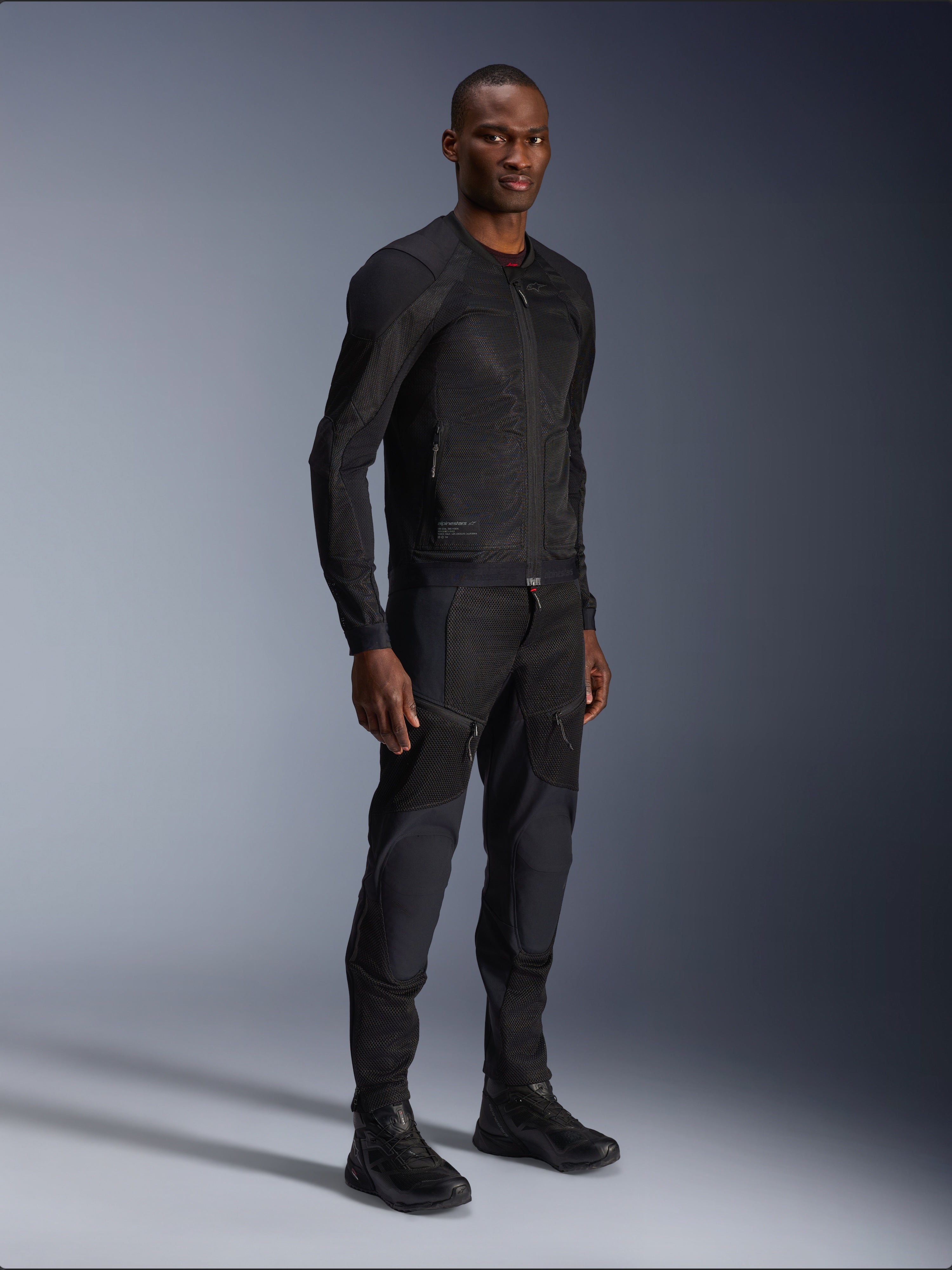 Alpinestars Troop-Air Jacke, Schwarz, technische Mesh-Motorradjacke getragen von einem Model mit passender schwarzer Hose und Stiefeln, leichte Mesh-Konstruktion, minimalistisches urbanes Design