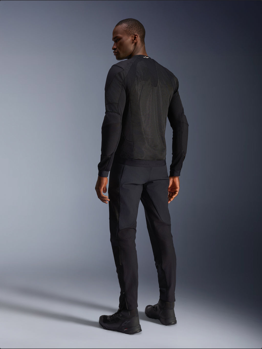 Alpinestars Troop-Air Jacke, schwarze technische Motorradjacke, Rückansicht mit großen Mesh-Einsätzen für erstklassige Belüftung, getragen mit passender schwarzer Hose und Motorradstiefeln, modernes urbanes Design für sommerliche Fahrten.