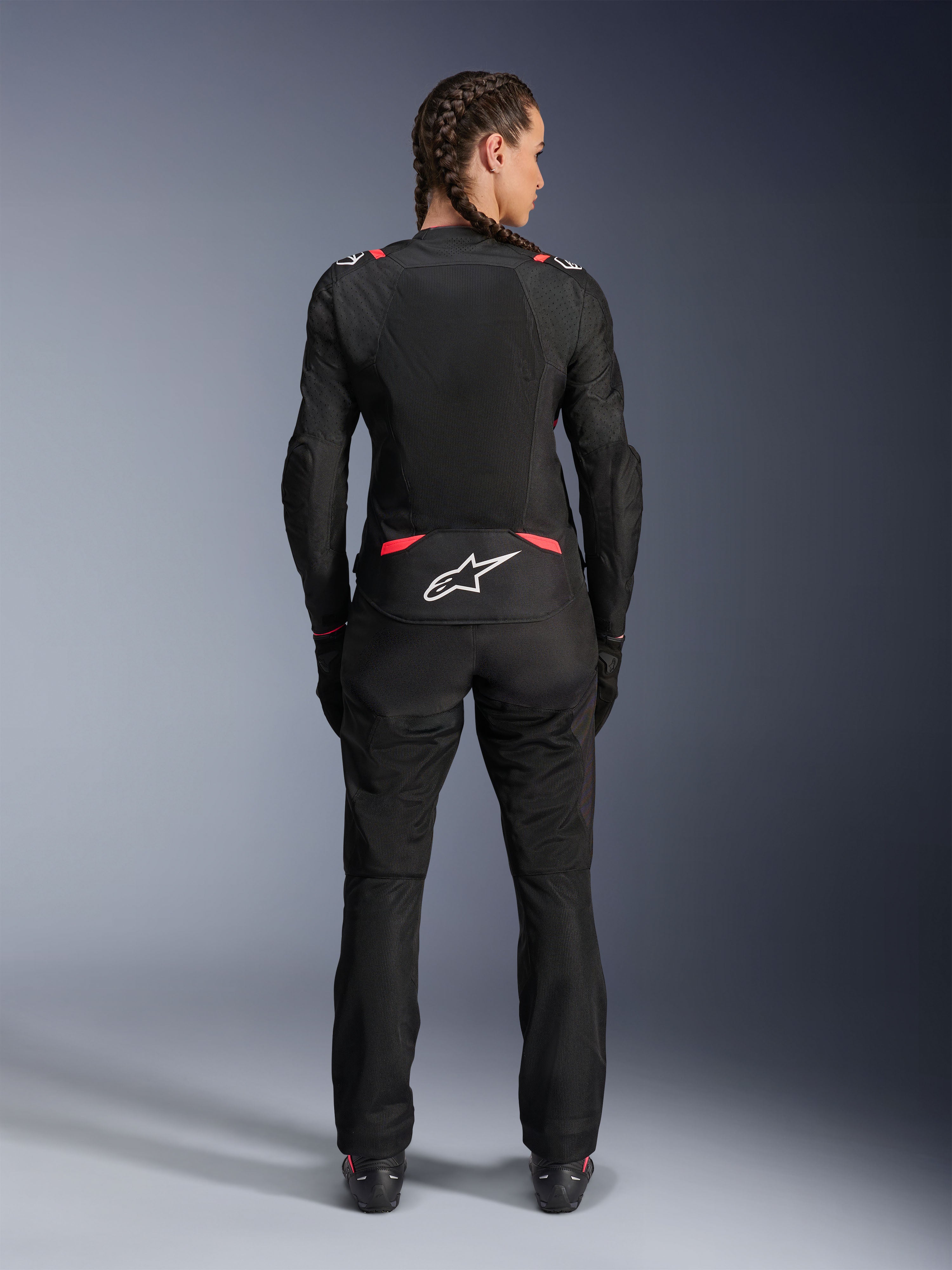 Alpinestars Stella T-SPS Air V2 Jacke, Damen-Textilmotorradjacke, Schwarz und Diva Pink, Rückansicht an einem Modell mit geflochtenem Haar, mit großen Mesh-Paneelen zur Belüftung, weißes Astars-Logo am unteren Rücken, kombiniert mit passender schwarzer Motorradhose.