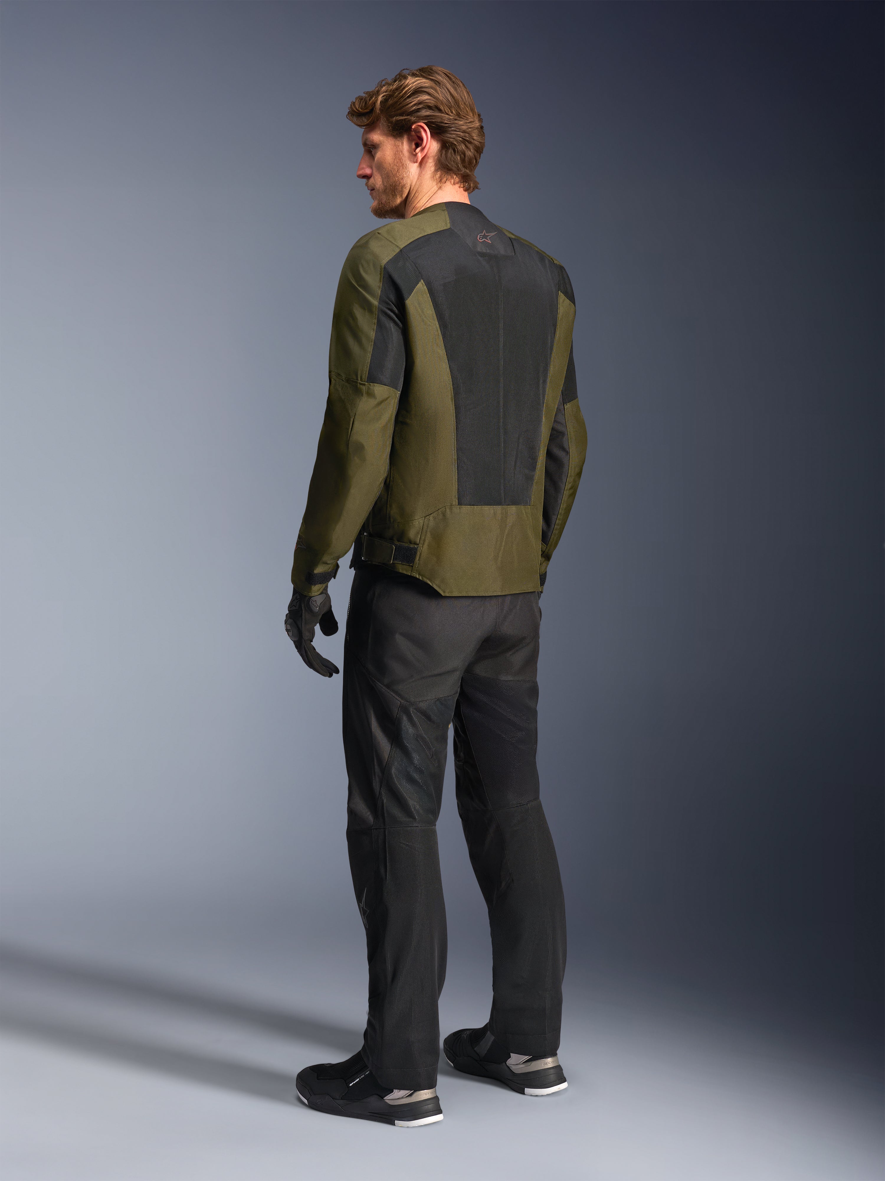 Männliches Model in Alpinestars C-1 Air Jacke in Khaki, urbane Motorradjacke, Rückansicht mit Fokus auf große schwarze Mesh-Paneele am Rücken, kombiniert mit schwarzer technischer Fahrhose und Handschuhen, moderne Motorradausrüstung für warmes Wetter