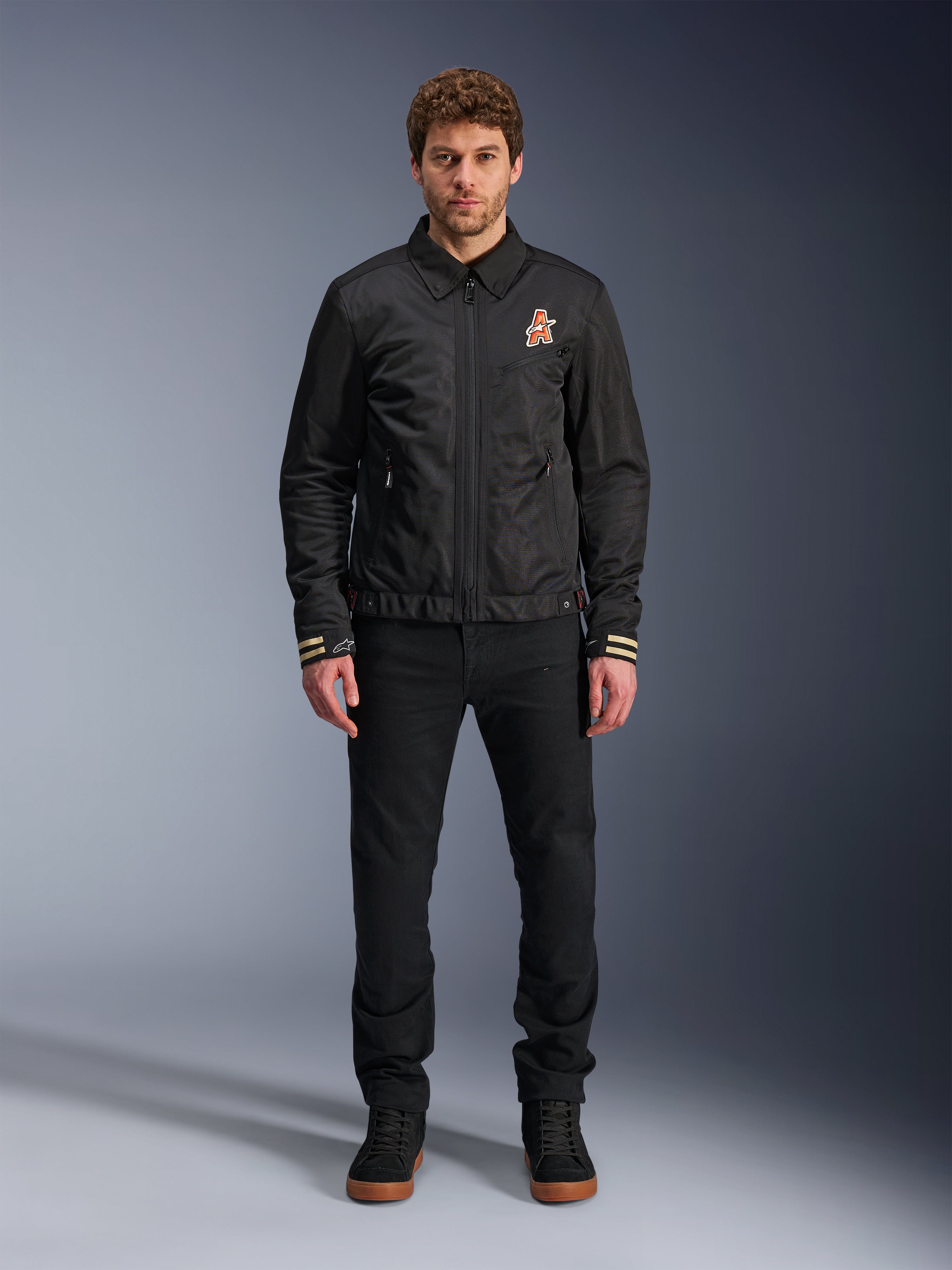 Männliches Model in der Alpinestars Ignite Air Jacke in Schwarz, mit klassischem Hemdkragen, Frontreißverschluss und einem orangefarbenen Logo auf der Brust. Kombiniert mit einer schwarzen Hose und schwarzen High-Top-Sneakern für einen modernen, urbanen Motorrad-Look.