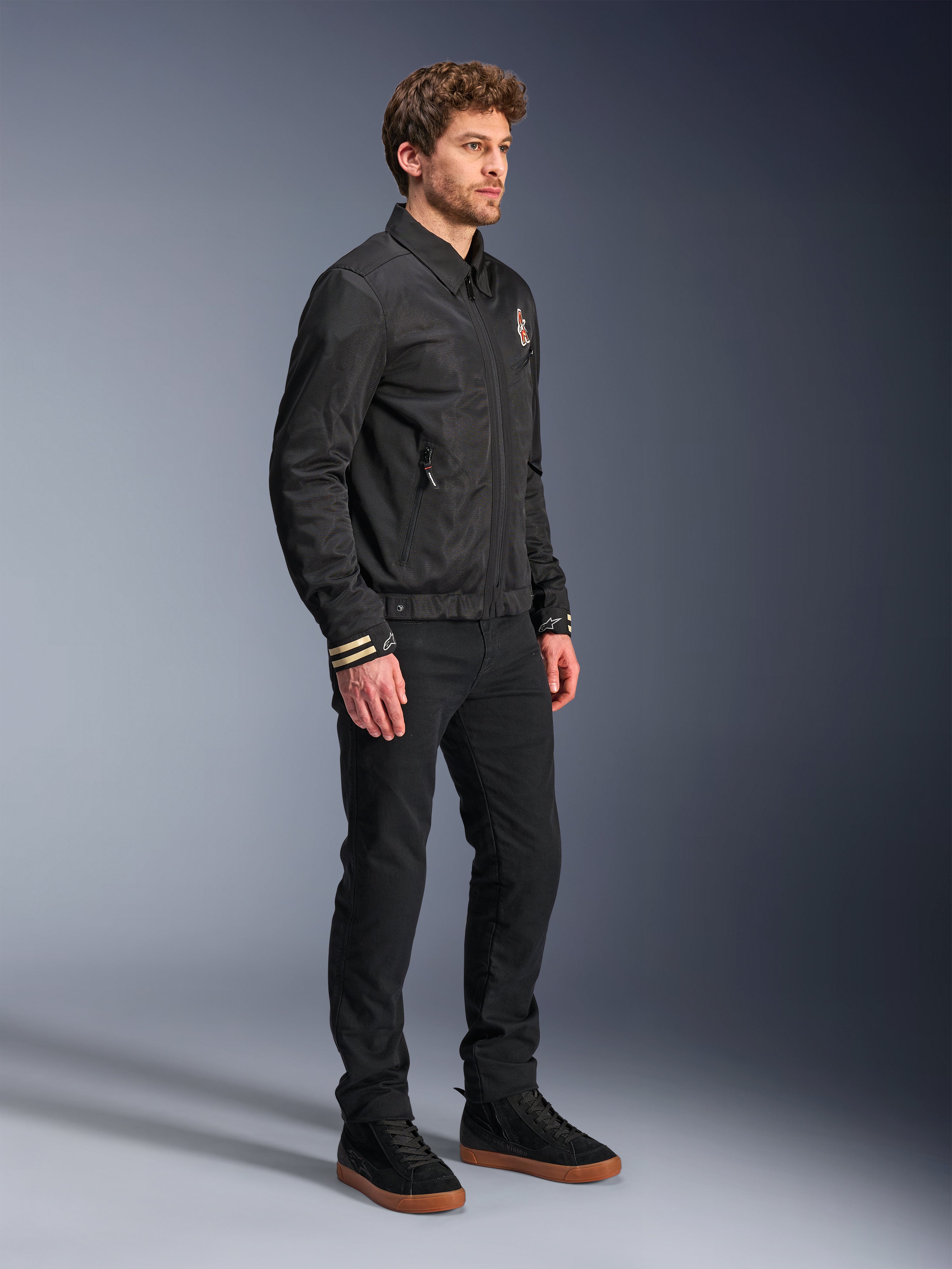 Alpinestars Ignite Air Jacke, Urbane Motorradjacke, Schwarz, getragen von einem Modell mit schwarzer Hose und schwarzen Sneakern, mit traditionellem Hemdkragen, Club-inspirierten Grafiken auf der Brust und an den Ärmelbündchen, elegantes, zeitgemäßes Design mit Mesh- und Satin-Finish