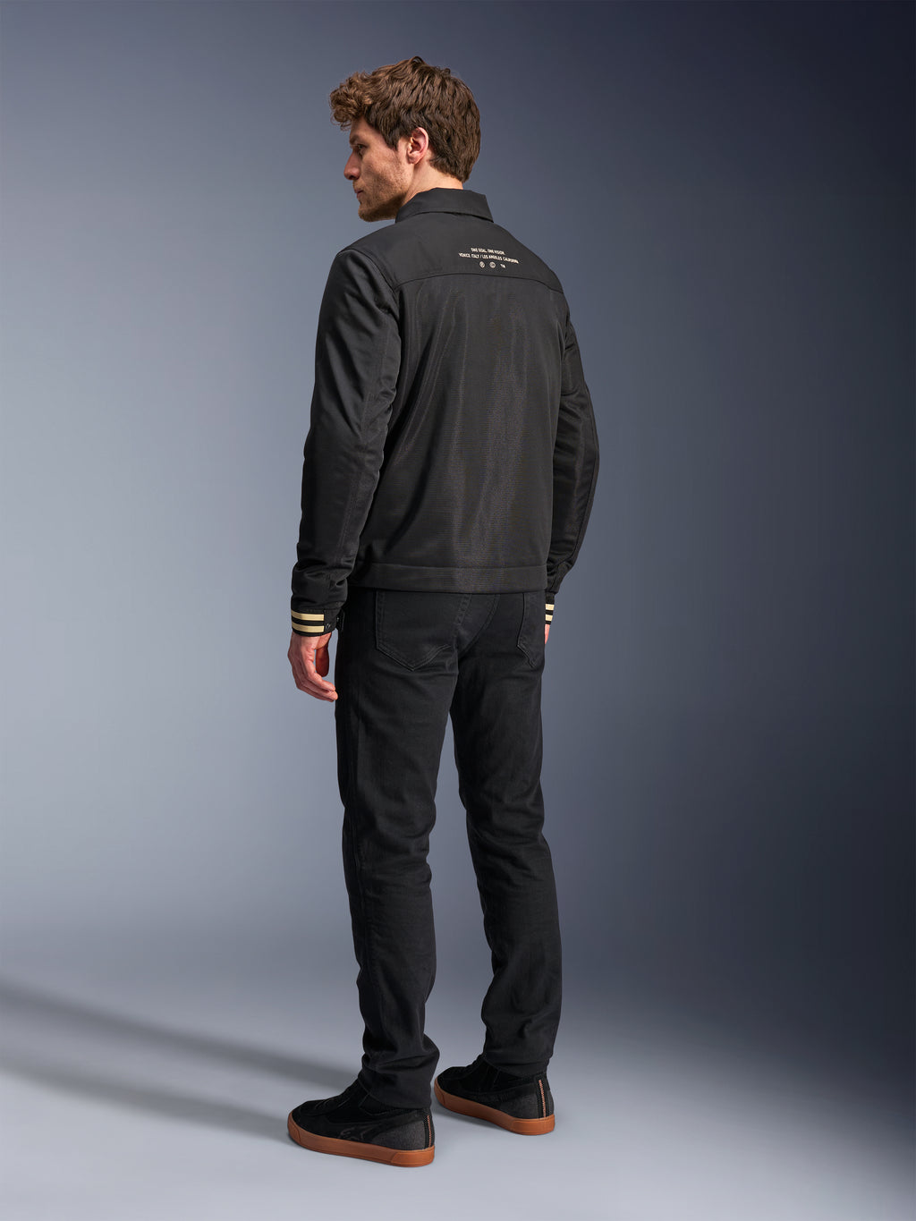 Alpinestars Ignite Air Jacke, schwarze Motorradjacke, Rückansicht mit weißem Grafiktext auf dem oberen Rücken und hellbraun gestreiften Bündchen, Silhouette im Harrington-Stil mit Mesh-Gewebe, kombiniert mit schwarzer Hose und schwarzen Sneakern