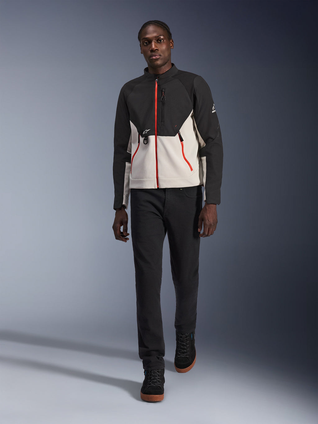 Männliches Model in der Alpinestars Provoke Jacke, urbane Motorradjacke in Schwarz und Metal Gray mit roten Reißverschluss-Akzenten, kombiniert mit schwarzer Hose und schwarzen High-Top-Schuhen, moderne Schutzausrüstung für Fahrten in der Stadt