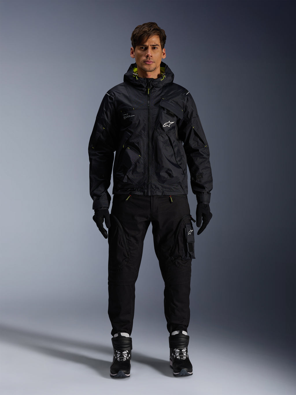 Männliches Model trägt die Alpinestars Mogress Air Jacke, schwarze Textiljacke mit Kapuze und Akzenten in Lime Punch, kombiniert mit einer schwarzen Utility-Hose, schwarzen Handschuhen und schwarz-weißen Motorradstiefeln, urbanes, vom Motorsport inspiriertes Streetwear-Design