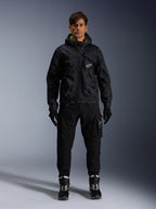Männliches Model trägt die Alpinestars Mogress Air Jacke, schwarze Textiljacke mit Kapuze und Akzenten in Lime Punch, kombiniert mit einer schwarzen Utility-Hose, schwarzen Handschuhen und schwarz-weißen Motorradstiefeln, urbanes, vom Motorsport inspiriertes Streetwear-Design