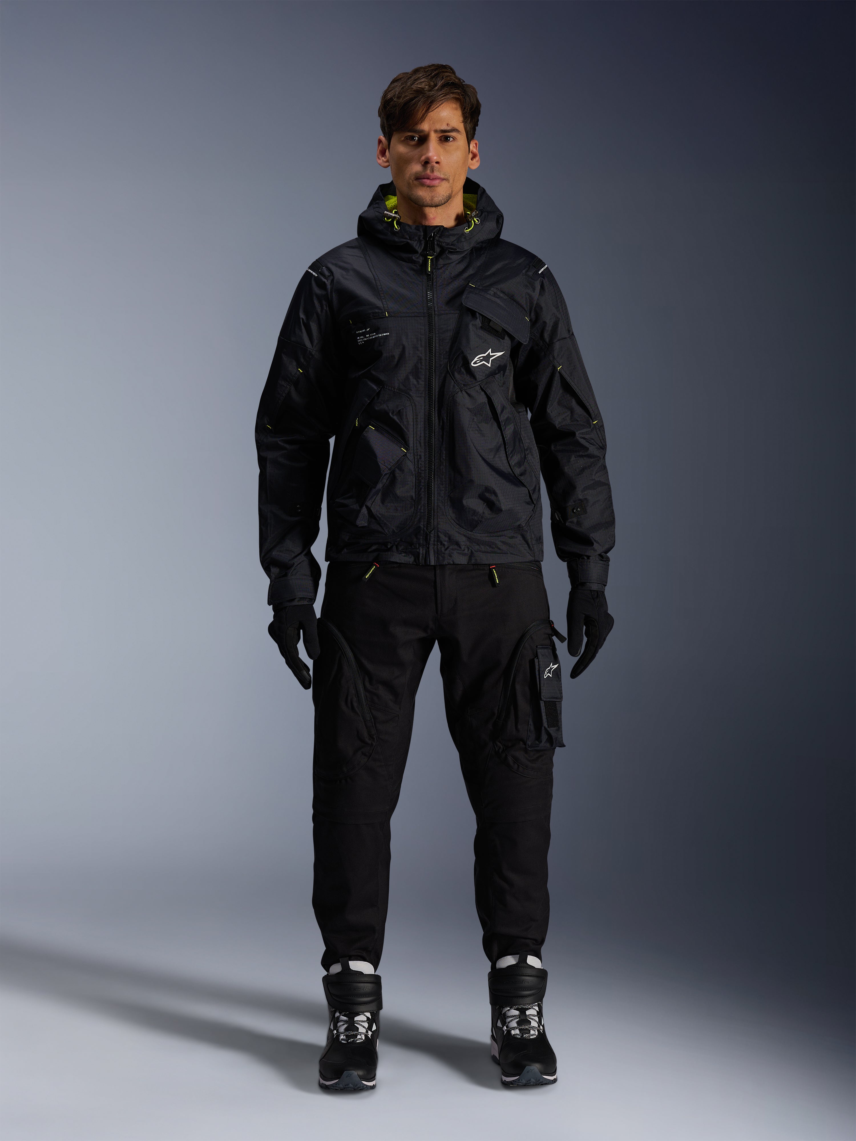 Männliches Model trägt die Alpinestars Mogress Air Jacke, schwarze Textiljacke mit Kapuze und Akzenten in Lime Punch, kombiniert mit einer schwarzen Utility-Hose, schwarzen Handschuhen und schwarz-weißen Motorradstiefeln, urbanes, vom Motorsport inspiriertes Streetwear-Design
