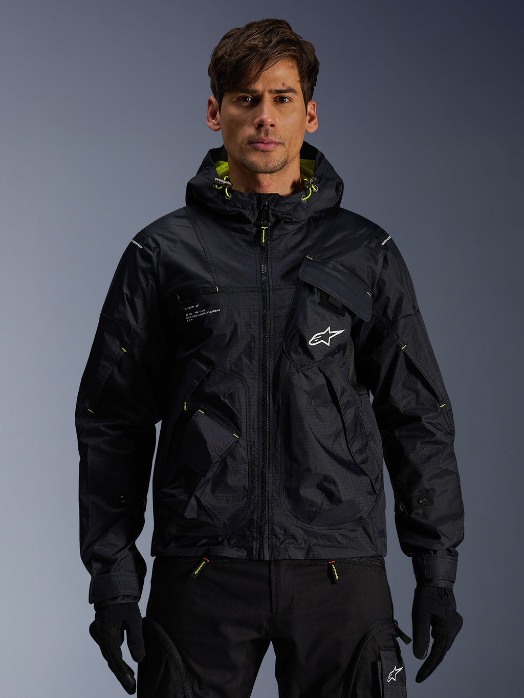 Alpinestars Mogress Air Jacke, urbane Motorradjacke, Schwarz mit Lime Punch-Akzenten, getragen von einem Model mit schwarzer Motorradhose und Handschuhen, Utility-inspiriertes Design mit Kapuze und weißem Stern-Logo auf der Brust, leichte Textilausrüstung für den Stadtverkehr