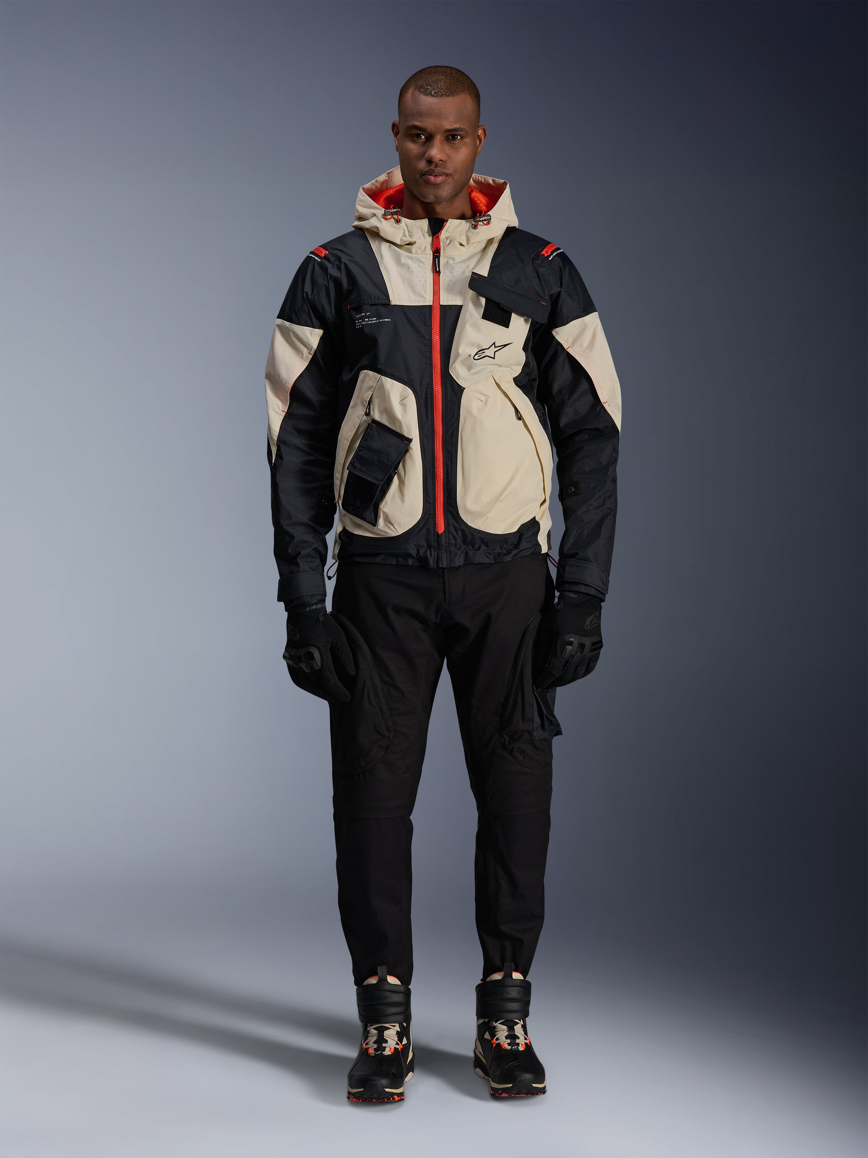 Alpinestars Mogress Air Jacke, urbane Motorradjacke, Schwarz, Almond und Hot Orange, getragen von einem männlichen Model mit schwarzer Hose und Handschuhen, Utility-inspiriertes Design mit cremefarbenen Einsätzen und leuchtend orangen Reißverschlüssen, moderne vom Motorsport beeinflusste Streetwear
