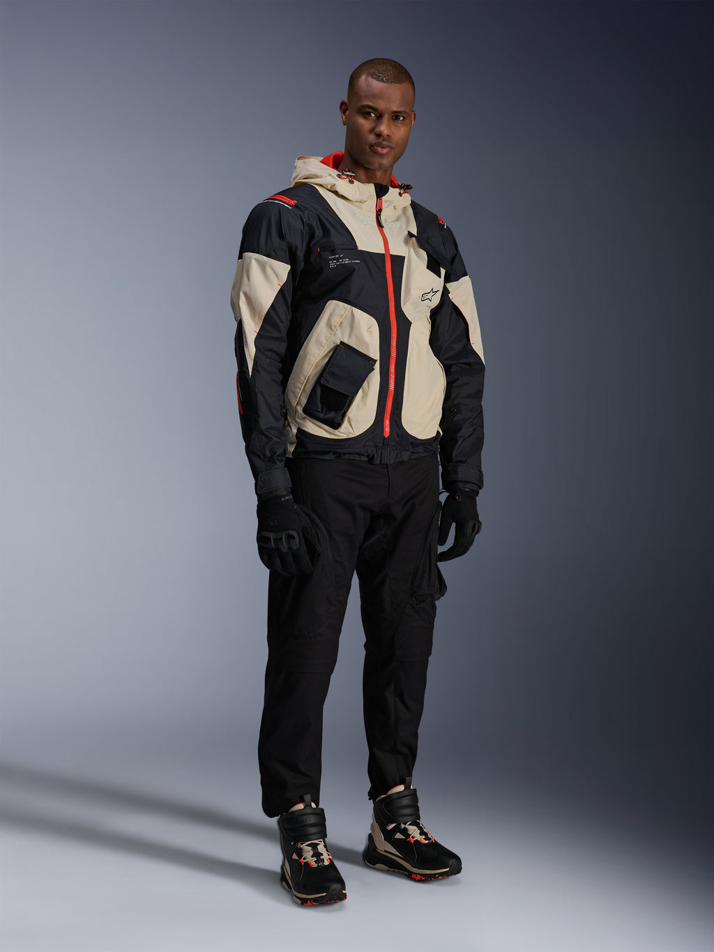 Model trägt die Alpinestars Mogress Air Jacke in Schwarz, Almond und Hot Orange, urbane Motorradjacke mit Utility-inspiriertem Design, kombiniert mit schwarzer Motorradhose, schwarzen Handschuhen und passenden technischen Stiefeln, moderne straßentaugliche Motorradausrüstung