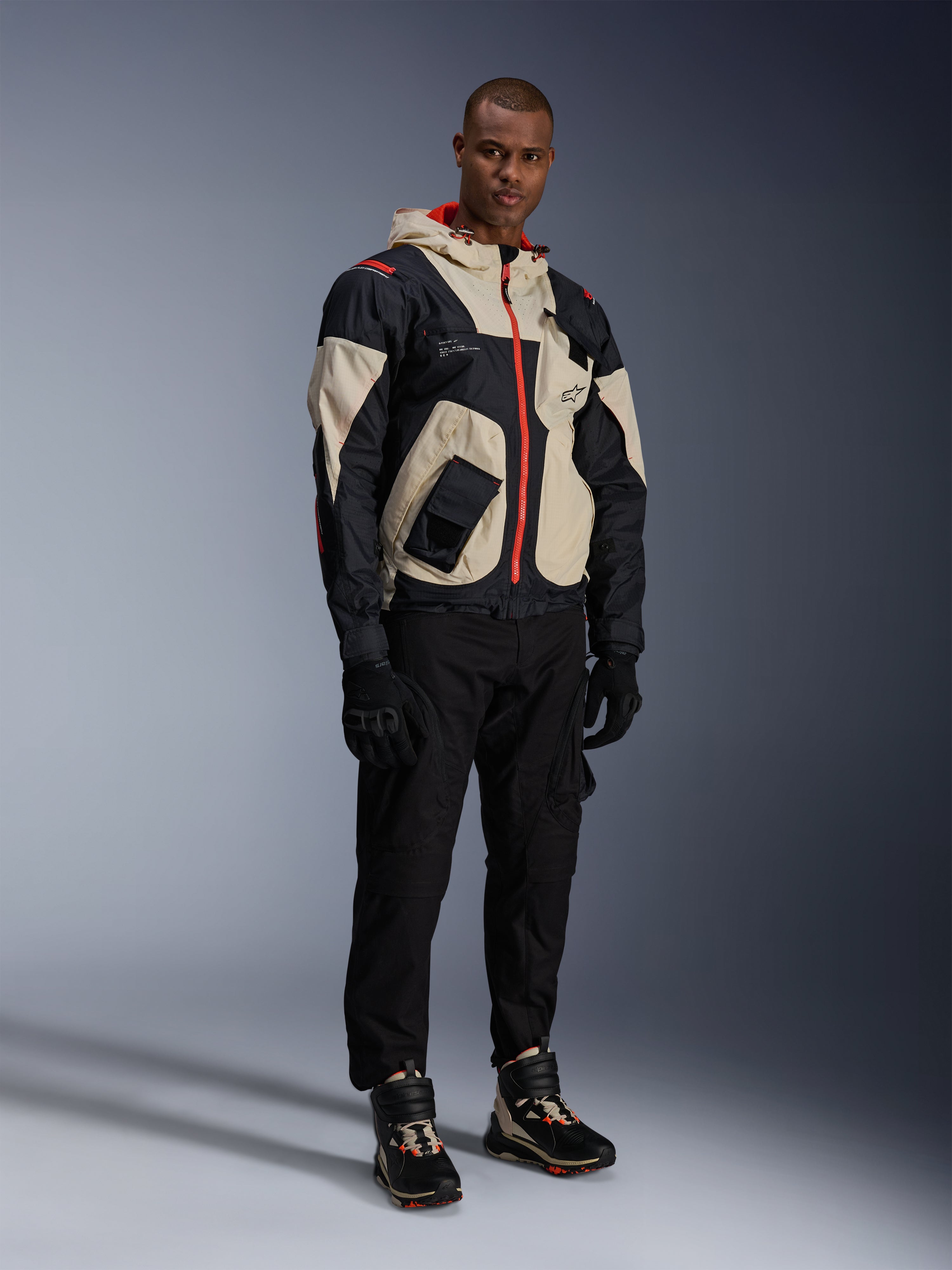 Model trägt die Alpinestars Mogress Air Jacke in Schwarz, Almond und Hot Orange, urbane Motorradjacke mit Utility-inspiriertem Design, kombiniert mit schwarzer Motorradhose, schwarzen Handschuhen und passenden technischen Stiefeln, moderne straßentaugliche Motorradausrüstung
