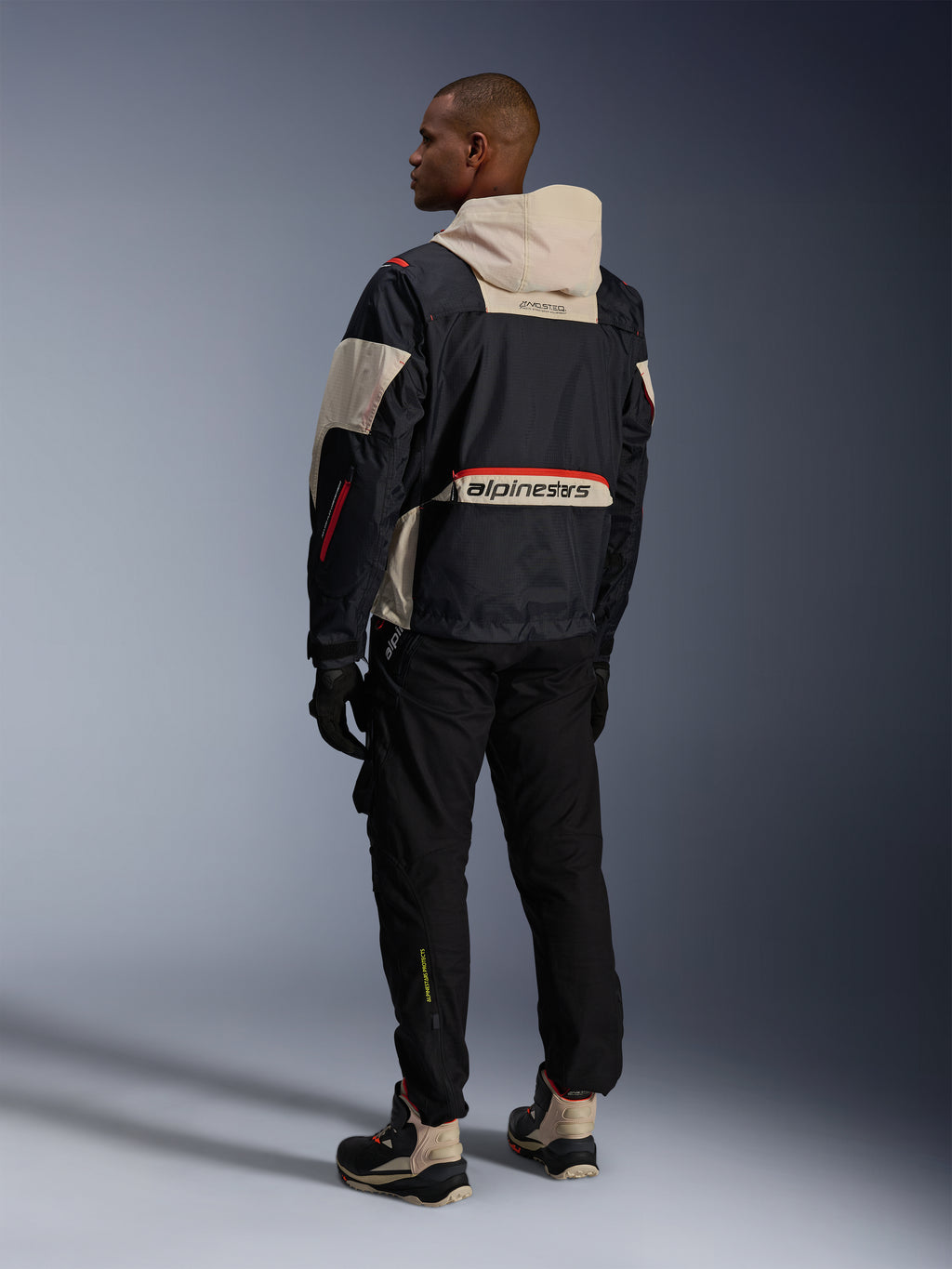 Alpinestars Mogress Air Jacke, Schwarz Mandel Hot Orange, Rückansicht eines Fahrers in urbaner Motorradausrüstung, mandelfarbene Kapuze und unteres Rückenpanel mit großem Alpinestars-Branding, orangefarbene Reißverschlussdetails, kombiniert mit schwarzer Hose und High-Top-Motorradstiefeln.