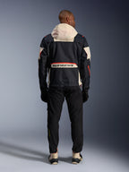 Rückansicht eines Fahrers in der Alpinestars Mogress Air Jacke in Schwarz, Almond und Hot Orange, mit einer cremefarbenen Kapuze, großem Alpinestars-Branding auf der Rückentasche und orangefarbenen Reißverschluss-Akzenten, kombiniert mit einer schwarzen Hose und cremefarbenen Stiefeln für einen urbanen Motorrad-Look.