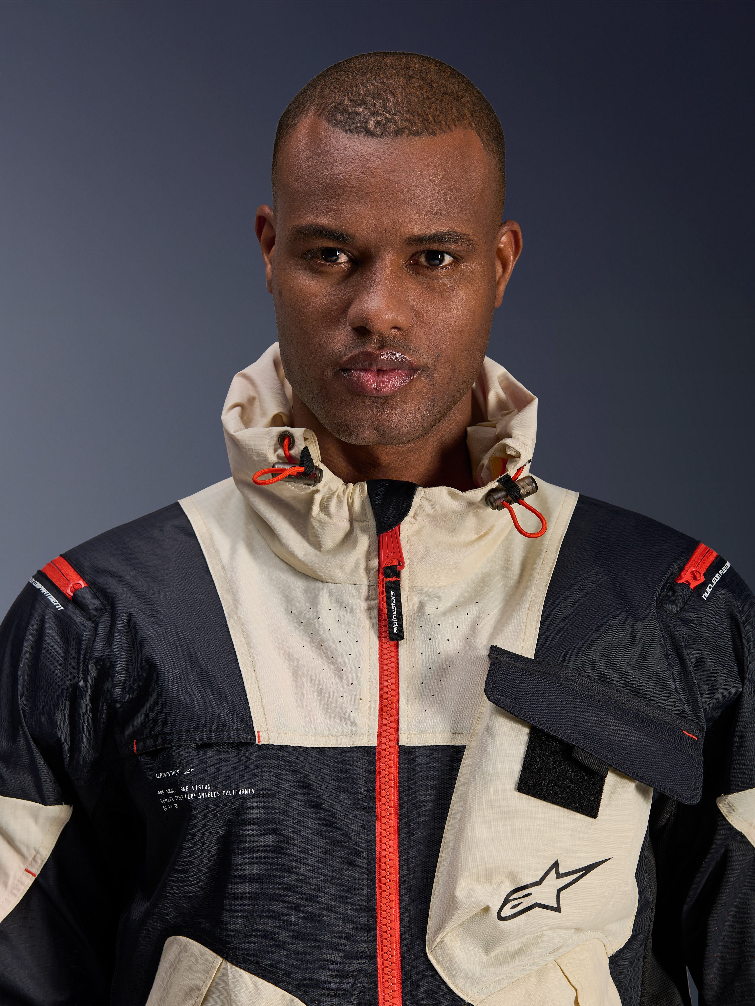 Männliches Model in der Alpinestars Mogress Air Jacke, urbane Motorradjacke, Schwarz, Mandelbeige und Hot Orange, Nahaufnahme des Color-Block-Designs, Ripstop-Gewebe, Kapuzenkragen mit orangefarbenen Kordelstoppern und Frontreißverschluss-Details.