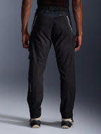 Alpinestars Acteon Hose, Urban-Motorradhose, Schwarz, Rückansicht mit verjüngter Passform, weißes Branding an den Oberschenkeln und gelbes Logo an der unteren Wade, verstellbare Beinabschlüsse mit Reißverschlüssen, kombiniert mit cremefarbenen Sneakern