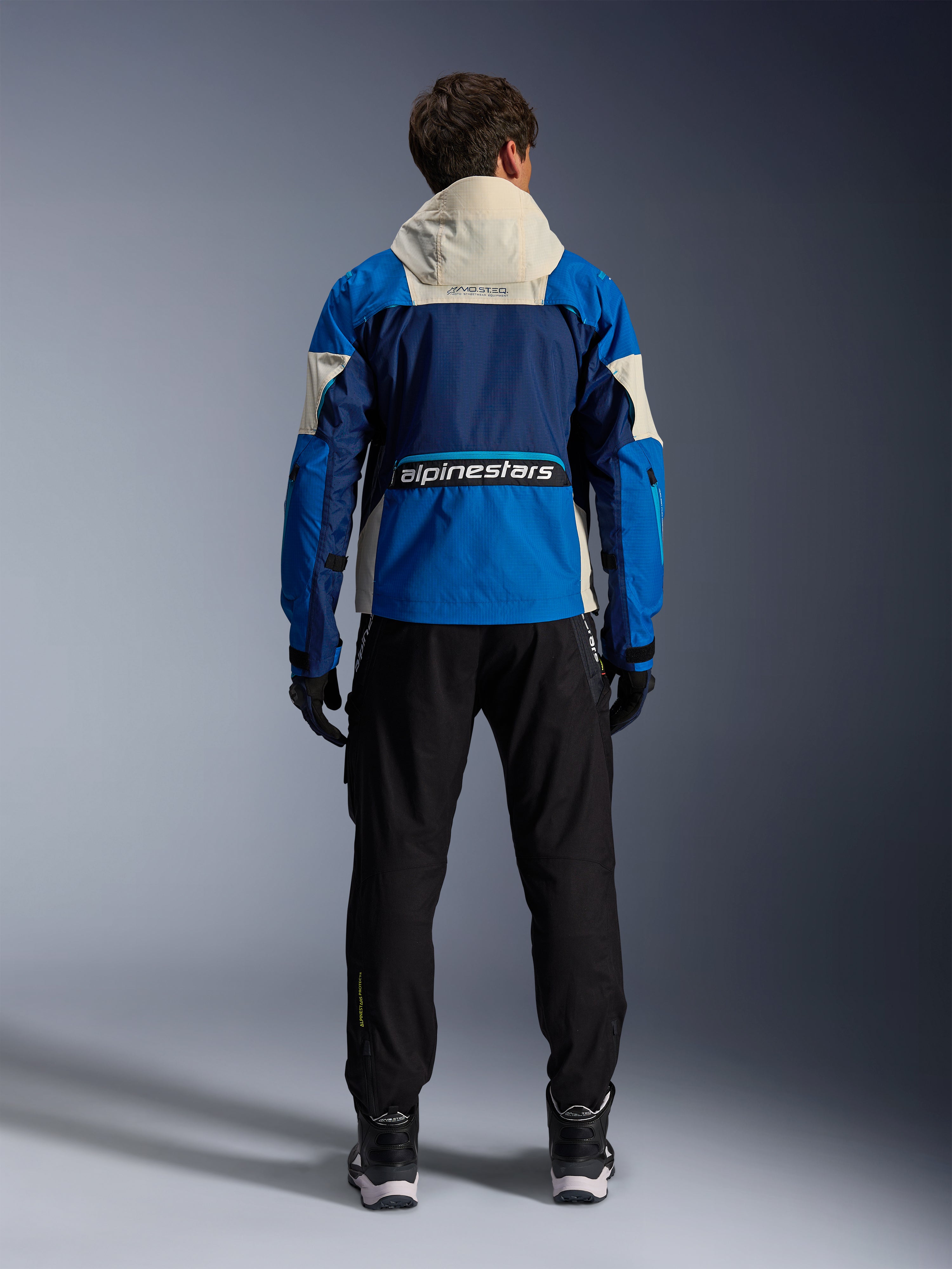 Alpinestars Mogress Air Jacke in Blue Almond, Rückansicht eines Fahrers in der Motorradjacke, mit navyfarbenen und hellblauen Einsätzen sowie einer cremeweißen Kapuze, markantes Alpinestars-Branding am unteren Rücken, urbaner, zeitgemäßer Stil für Motorradbekleidung