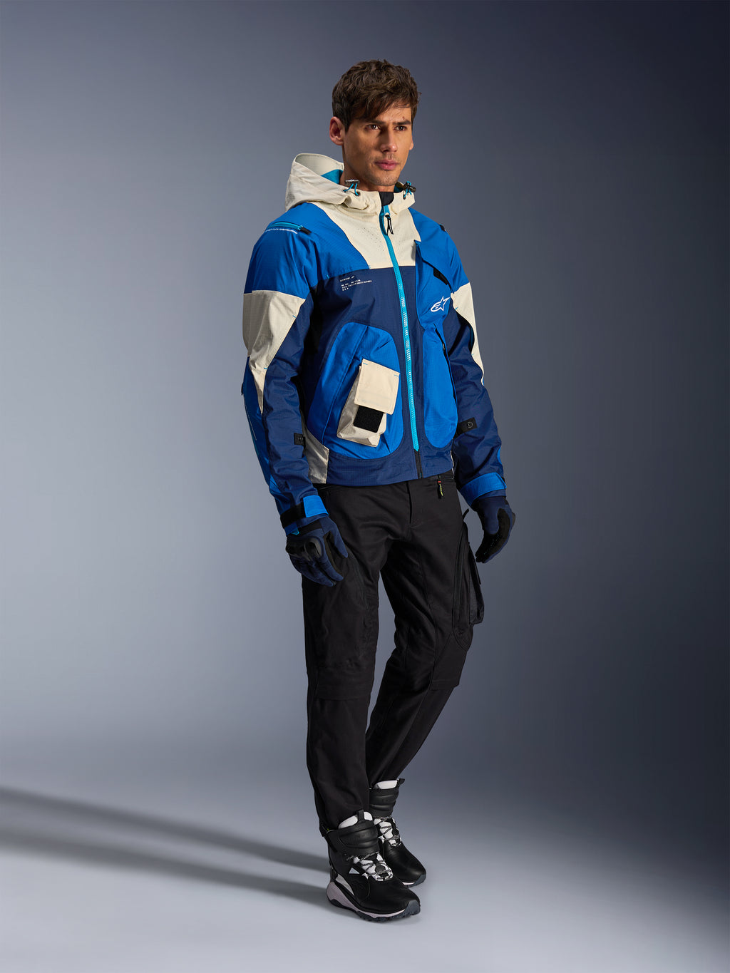 Alpinestars Mogress Air Jacke, urbane Motorradjacke, Blue Almond mit cremefarbenen und marineblauen Einsätzen, mit Kapuze und Utility-Taschen, getragen mit schwarzer Motorradhose, Handschuhen und Stiefeln, moderne, von Streetwear inspirierte Schutzausrüstung.