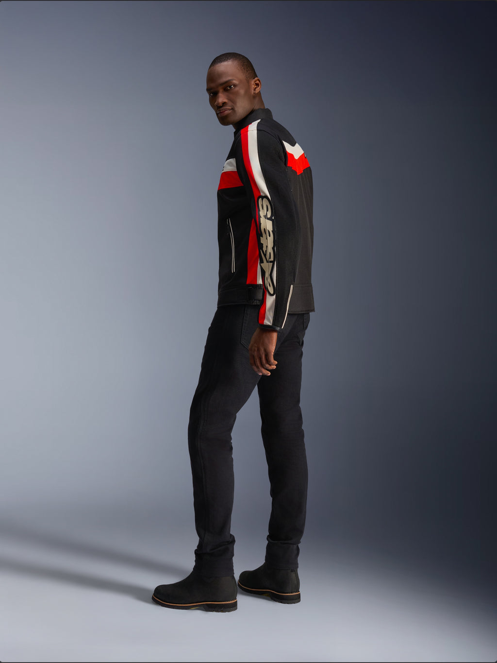 Ein Mann in der Alpinestars T-Dyno Air Jacke in Schwarz, Bright Red und Ecru, vintage-inspirierte Textil-Motorradjacke mit Mesh-Einsätzen, markantes Branding auf dem Ärmel, kombiniert mit schwarzer Hose und Stiefeln für einen modernen Biker-Look