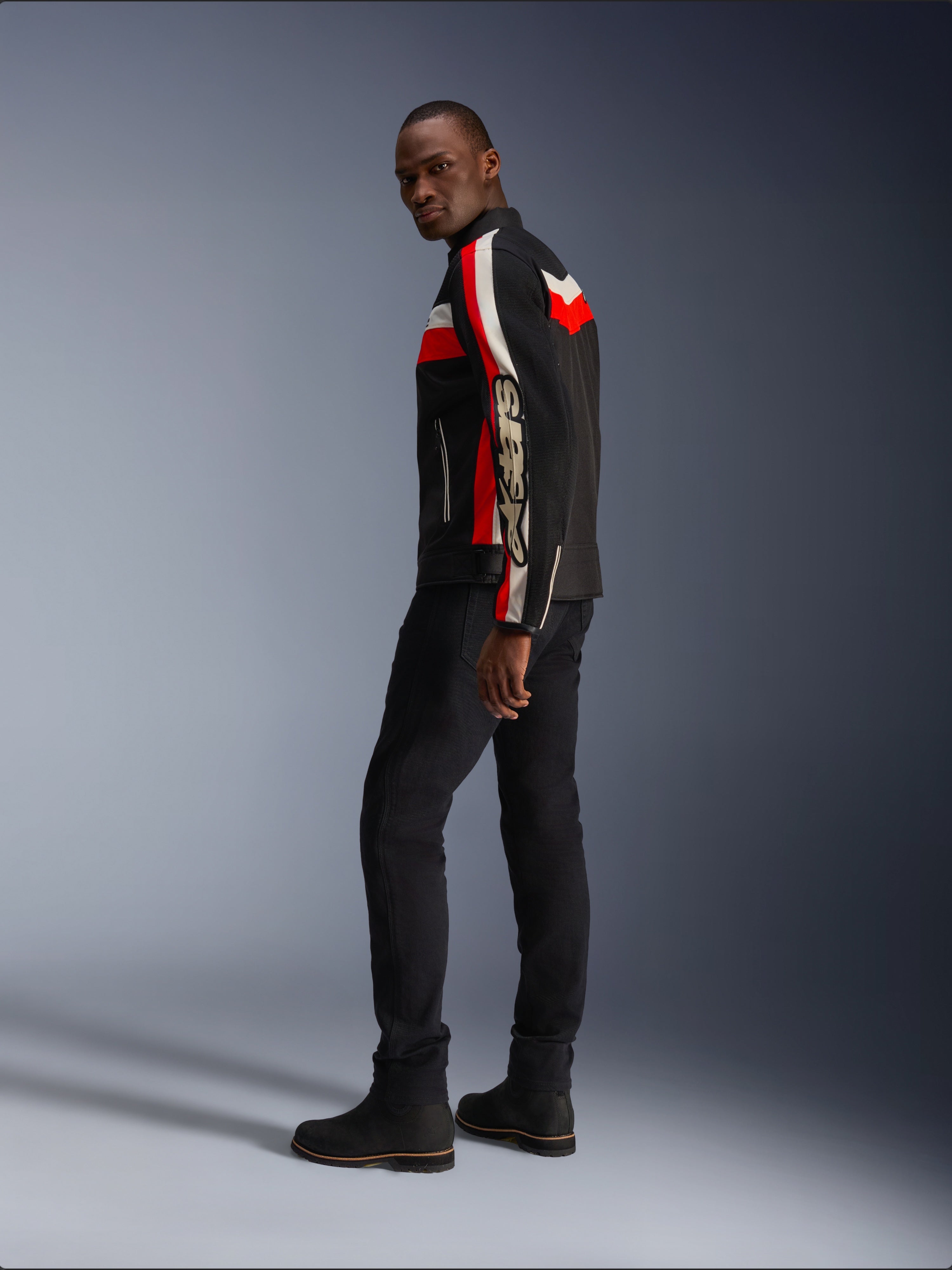 Ein Mann in der Alpinestars T-Dyno Air Jacke in Schwarz, Bright Red und Ecru, vintage-inspirierte Textil-Motorradjacke mit Mesh-Einsätzen, markantes Branding auf dem Ärmel, kombiniert mit schwarzer Hose und Stiefeln für einen modernen Biker-Look