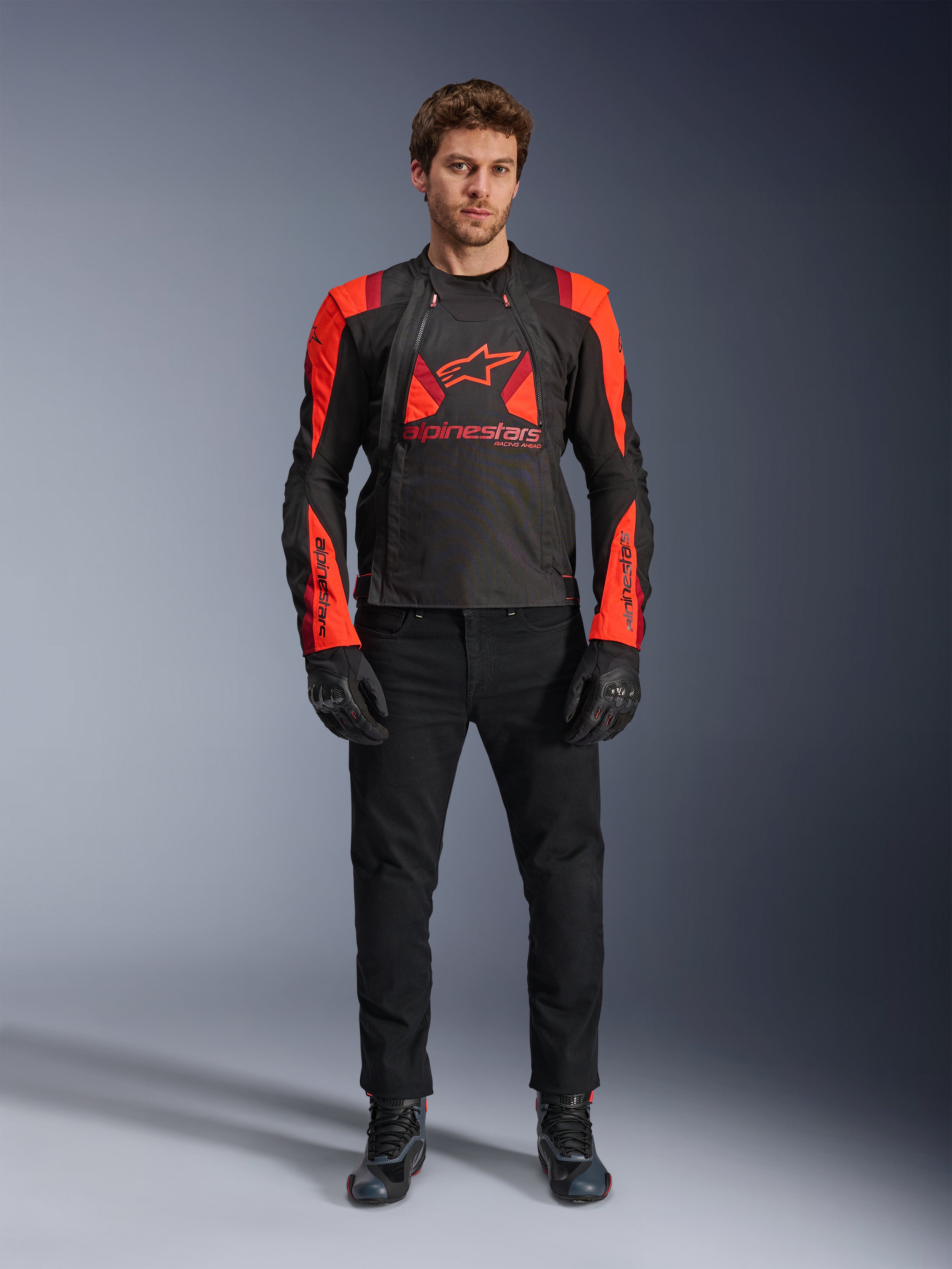 Alpinestars T-Stunt Air Jacke, Schwarz, Leuchtrot und Dunkelrot, getragen von einem Model mit schwarzen Motorradjeans und Handschuhen, mit modularem 2-in-1-Design und markanten Mesh-Einsätzen für Luftzirkulation, sportlich geschnittene Motorradausrüstung für die Straße
