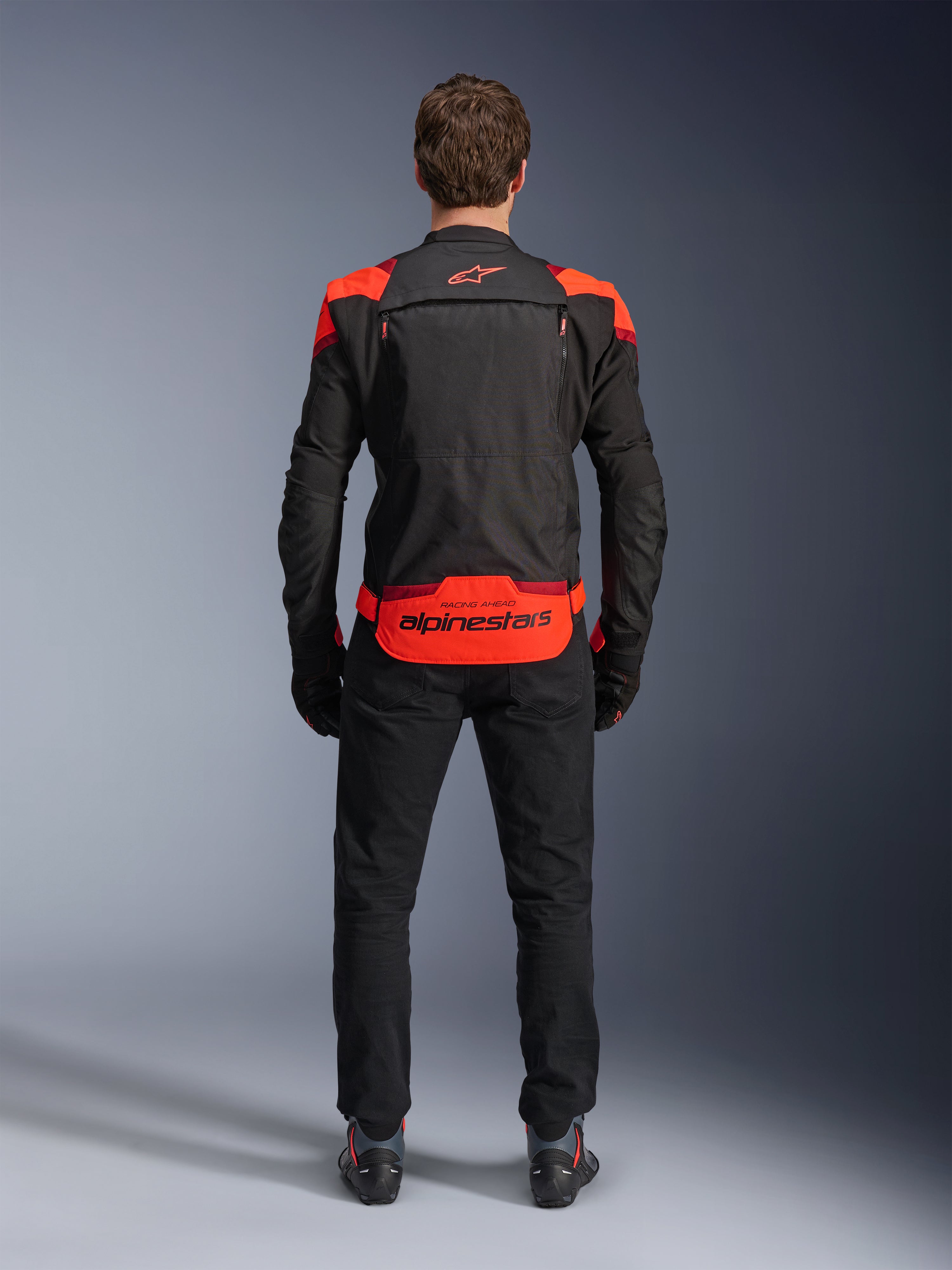 Alpinestars T-Stunt Air Jacke, Schwarz mit Red Fluo und dunkelroten Akzenten, Rückansicht am Model, kombiniert mit schwarzer Motorradhose und Stiefeln, markantes Alpinestars-Branding am unteren Rücken, modulares Design mit Mesh-Konstruktion für Belüftung