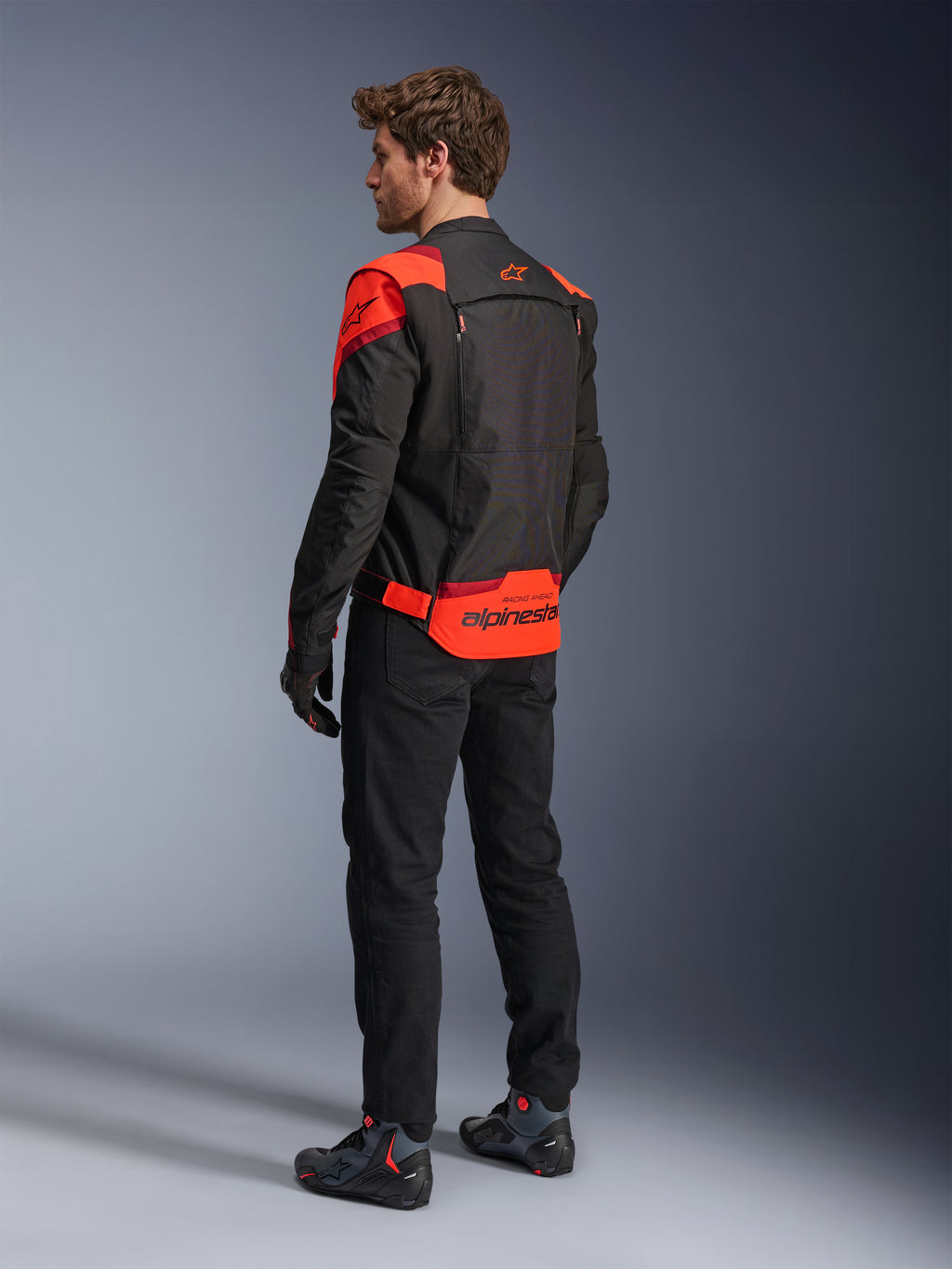 Mann in Alpinestars T-Stunt Air Jacke, Rückansicht, Schwarz, Fluo-Rot und Dunkelrot, Textil-Motorradjacke mit Mesh-Einsätzen und sportlichem Design, markantes Alpinestars-Branding auf dem unteren Rücken, kombiniert mit schwarzer Hose und Motorradstiefeln