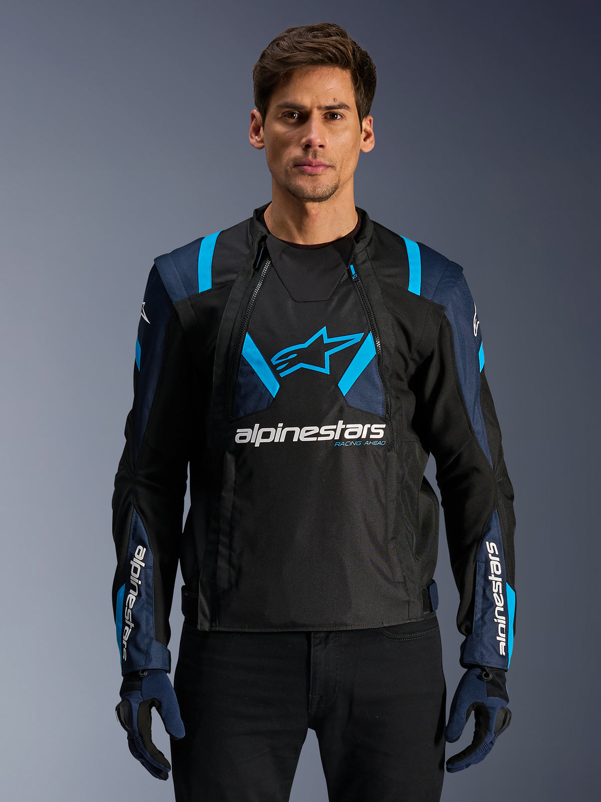 Alpinestars T-Stunt Air Jacke, Motorrad-Meshjacke, Schwarz und Blau, getragen von einem Model mit schwarzer Hose und Handschuhen, mit modularem 2-in-1-Design, atmungsaktiven Tech-Mesh-Einsätzen, Stern-Logo auf der Brust und Alpinestars-Branding auf den Ärmeln.