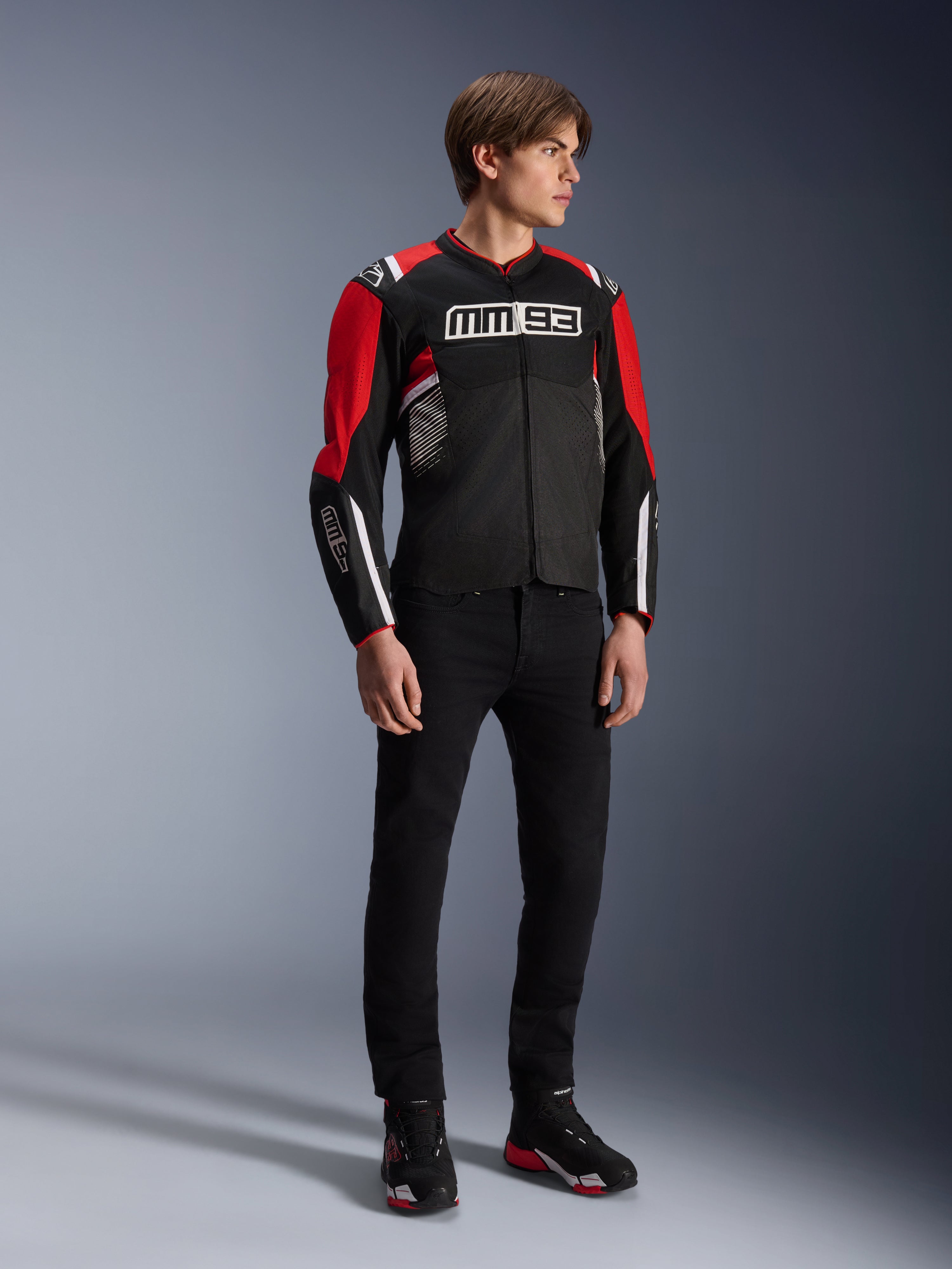 Male model wearing the Alpinestars MM93 T-SPS Air v2 Jacket in Black Bright Red White, featuring prominent MM93 branding on the chest and sleeves, paired with black pants and riding boots, sporty urban motorcycle style with visible mesh panels for airflow.