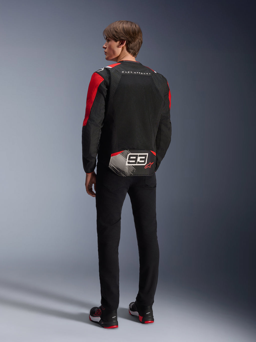 Model from behind wearing the Alpinestars MM93 T-SPS Air V2 Jacket, Black with Bright Red and White details, featuring 'MARC MARQUEZ' text and large '93' logo on the back tail, mesh textile construction for ventilation, worn with black pants and coordinated black and red riding shoes