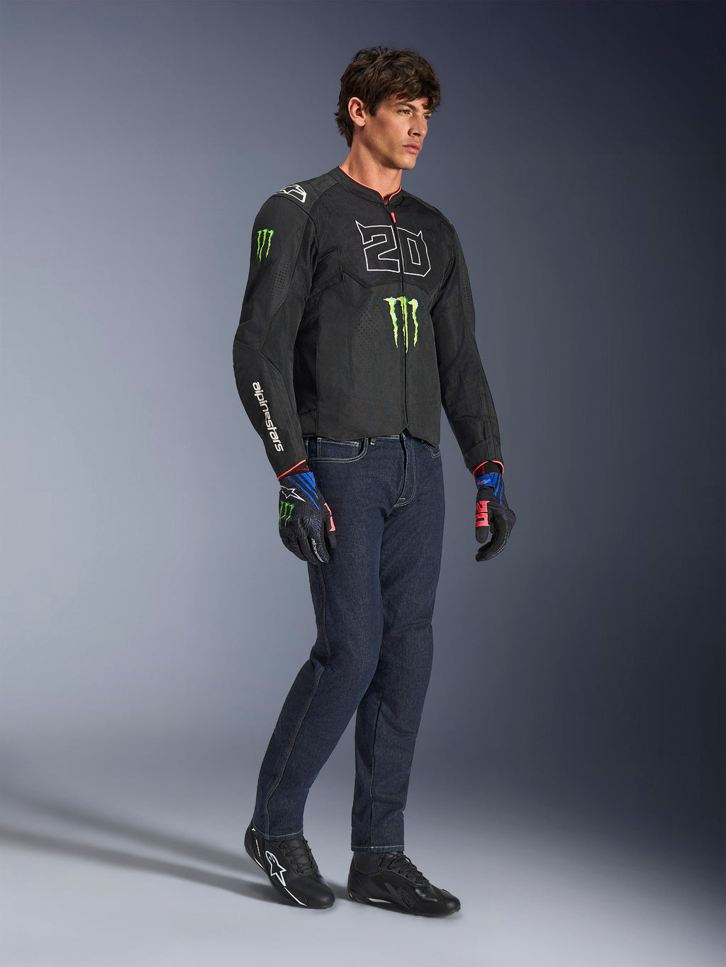 Male rider wearing the Alpinestars FQ20 T-SPS Air V2 Monster Jacket in black, featuring the '20' logo and Monster Energy claw on the chest, paired with dark riding jeans, black motorcycle shoes, and matching FQ20 gloves, full-body studio shot.