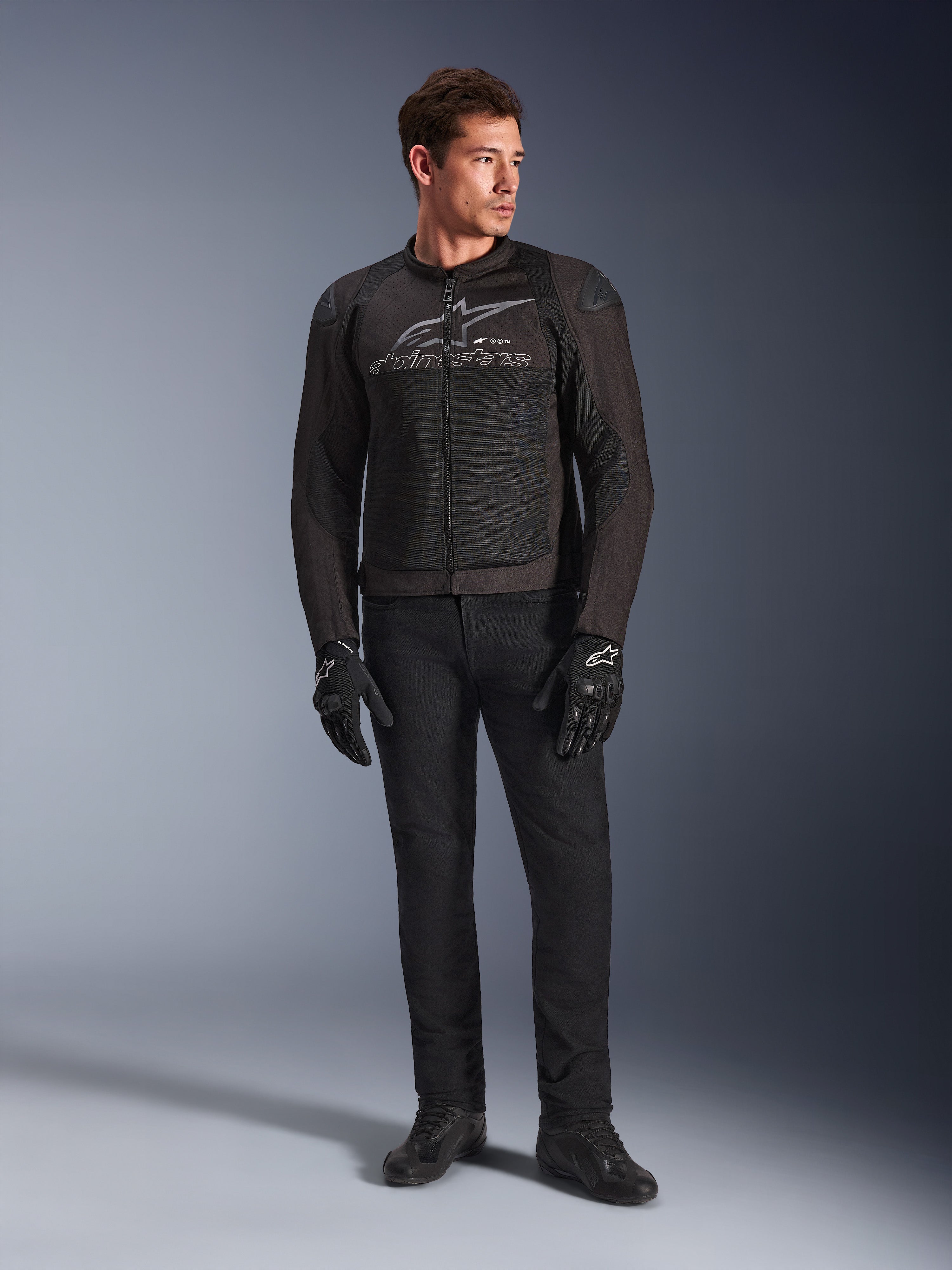 Male model wearing Alpinestars SMX Air Jacket, Black Anthracite motorcycle jacket with ventilated mesh panels and grey chest logo, paired with black pants and protective gloves, studio setting with a gradient background
