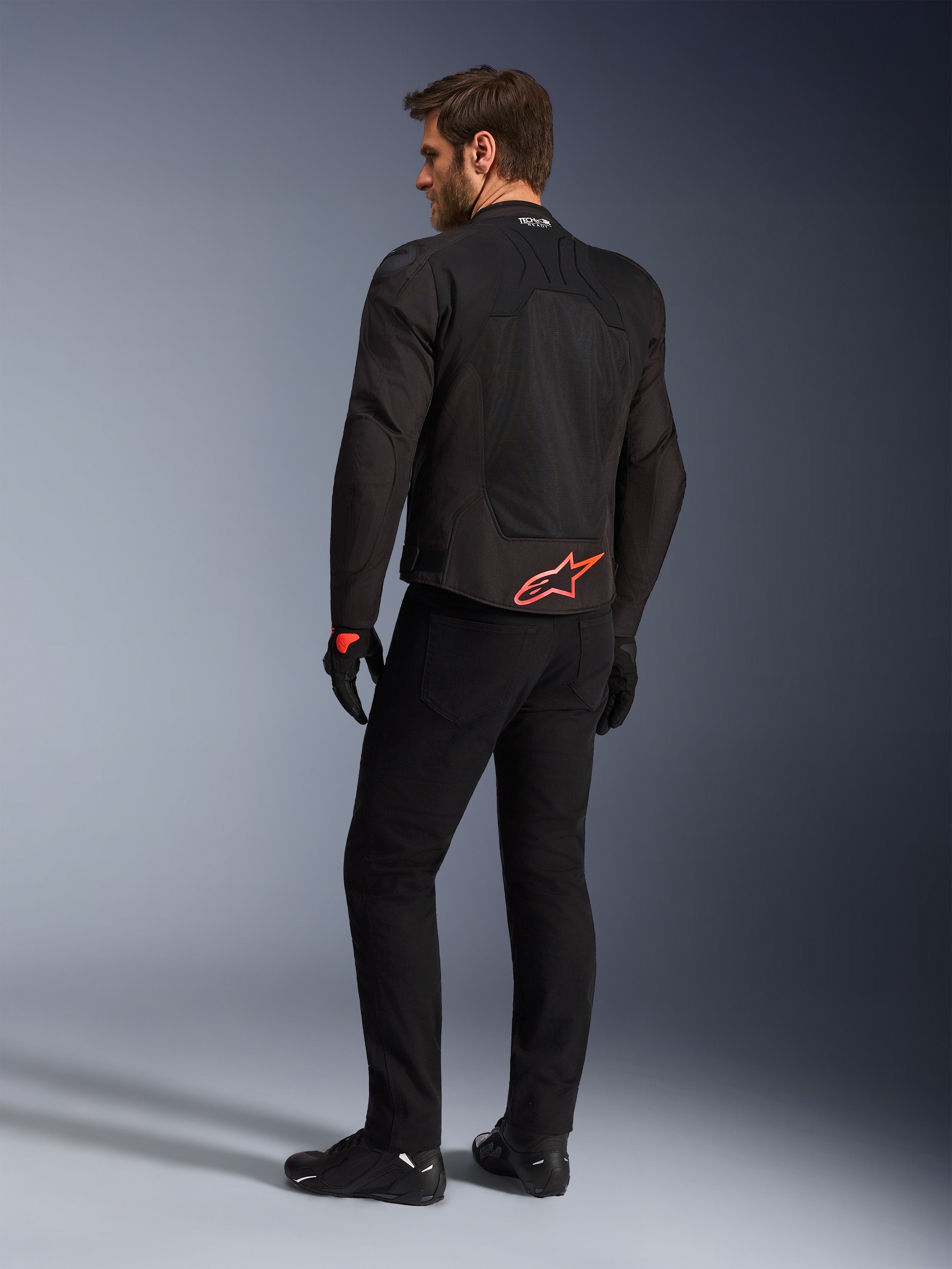 Rückansicht eines Modells in der Alpinestars SMX Air Jacke in Schwarz, mit einem leuchtend roten Alpinestars-Logo am unteren Rücken, „Tech-Air Ready“-Branding am Kragen und technischen Mesh-Einsätzen zur Belüftung, kombiniert mit schwarzer Motorradhose und Handschuhen.