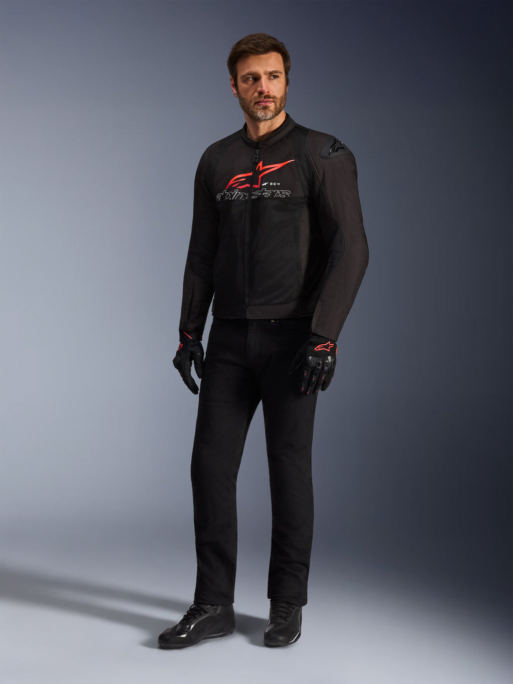 Männliches Model in der Alpinestars SMX Air Jacke in Schwarz Bright Red Weiß, kombiniert mit schwarzer technischer Hose, schwarz-roten Handschuhen und schwarzen Motorradschuhen, mit markantem rotem Brustlogo und weißem Branding, Ganzkörper-Studioaufnahme für Sport-Motorradausrüstung