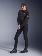 Weibliches Model in der Alpinestars Stella Grace Damenjacke, schwarze Urban-Motorradjacke, kombiniert mit schwarzer Motorradhose und Stiefeln, Workwear-inspiriertes Design mit bronzefarbenem gesticktem Logo auf der Brust und kreisförmigem Spiral-Aufnäher am Ärmel