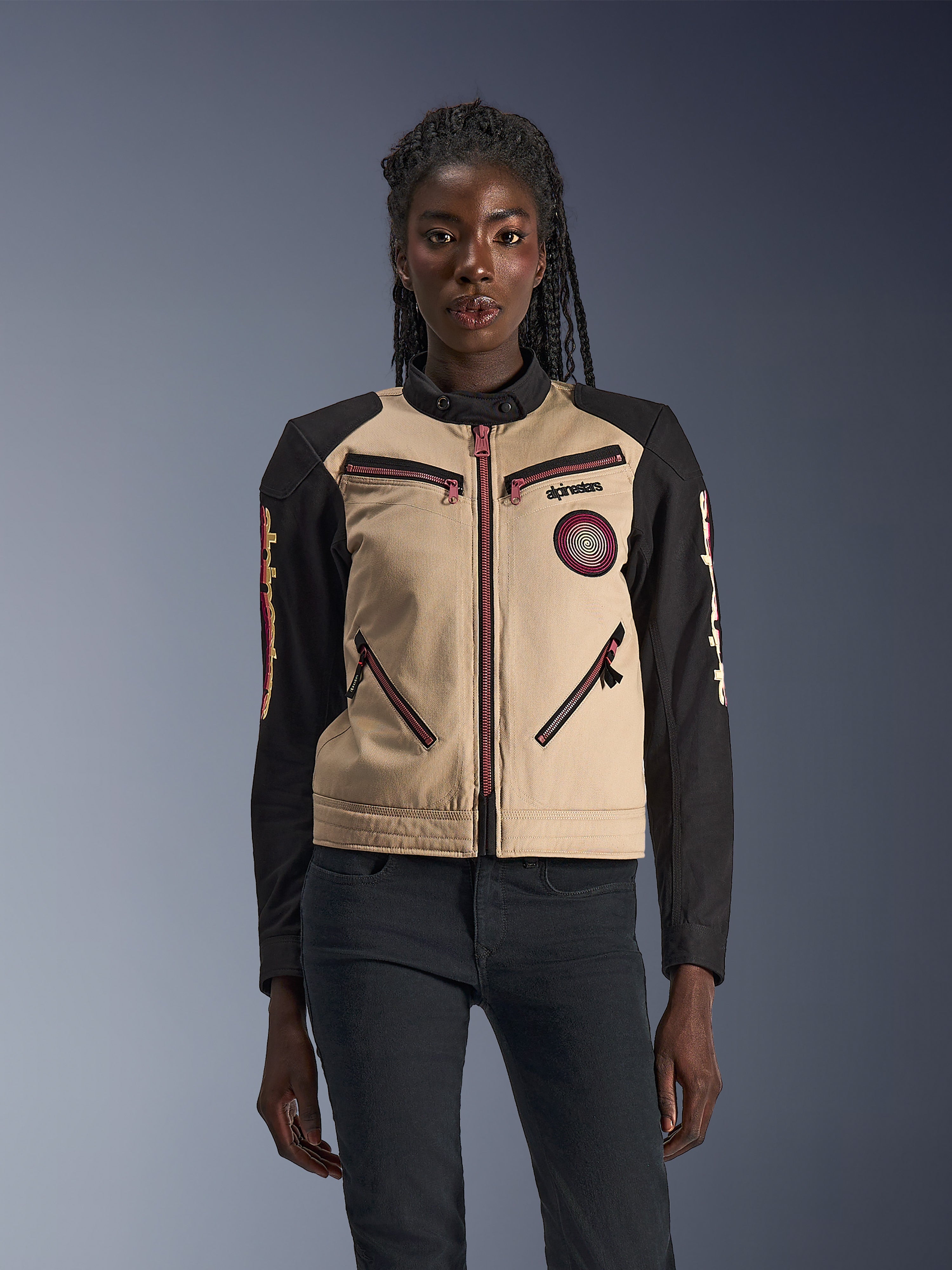 Alpinestars Stella Grace Jacke, Damen-Motorradjacke, Aluminum und Schwarz, mit einem von Workwear inspirierten Design, pink-roten Reißverschluss-Akzenten, einem spiralförmigen Brust-Aufnäher und langen schwarzen Ärmeln mit gelben und roten Grafiken, getragen von einem Model.