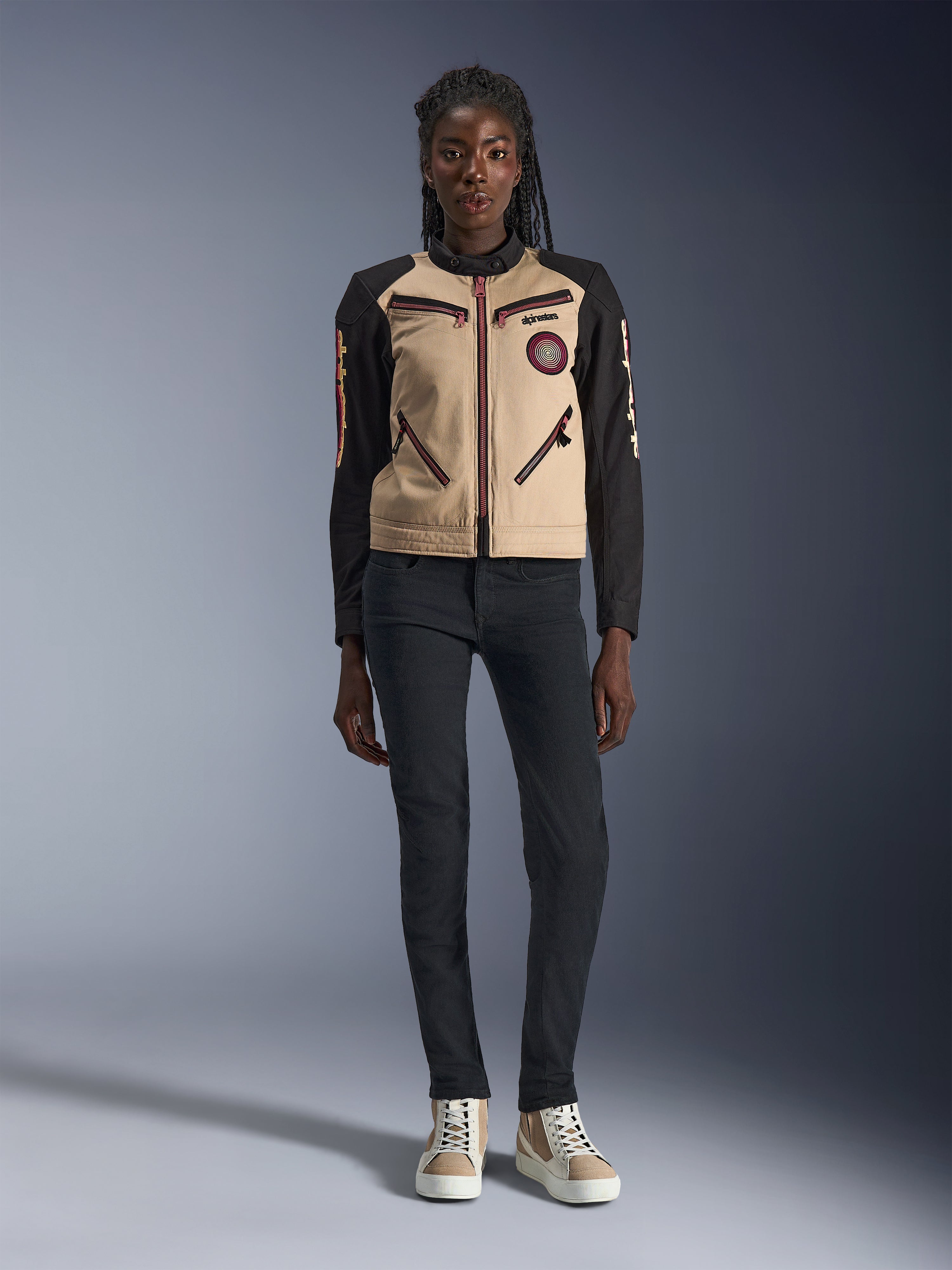 Alpinestars Stella Grace Damenjacke, urbane Motorradjacke, hellbraune und schwarze Textilkonstruktion, mit Reißverschlüssen vorne und besticktem Oscar-Spiral-Aufnäher, getragen mit schwarzer Hose und High-Top-Sneakern, moderne, von Workwear inspirierte Fahrausrüstung