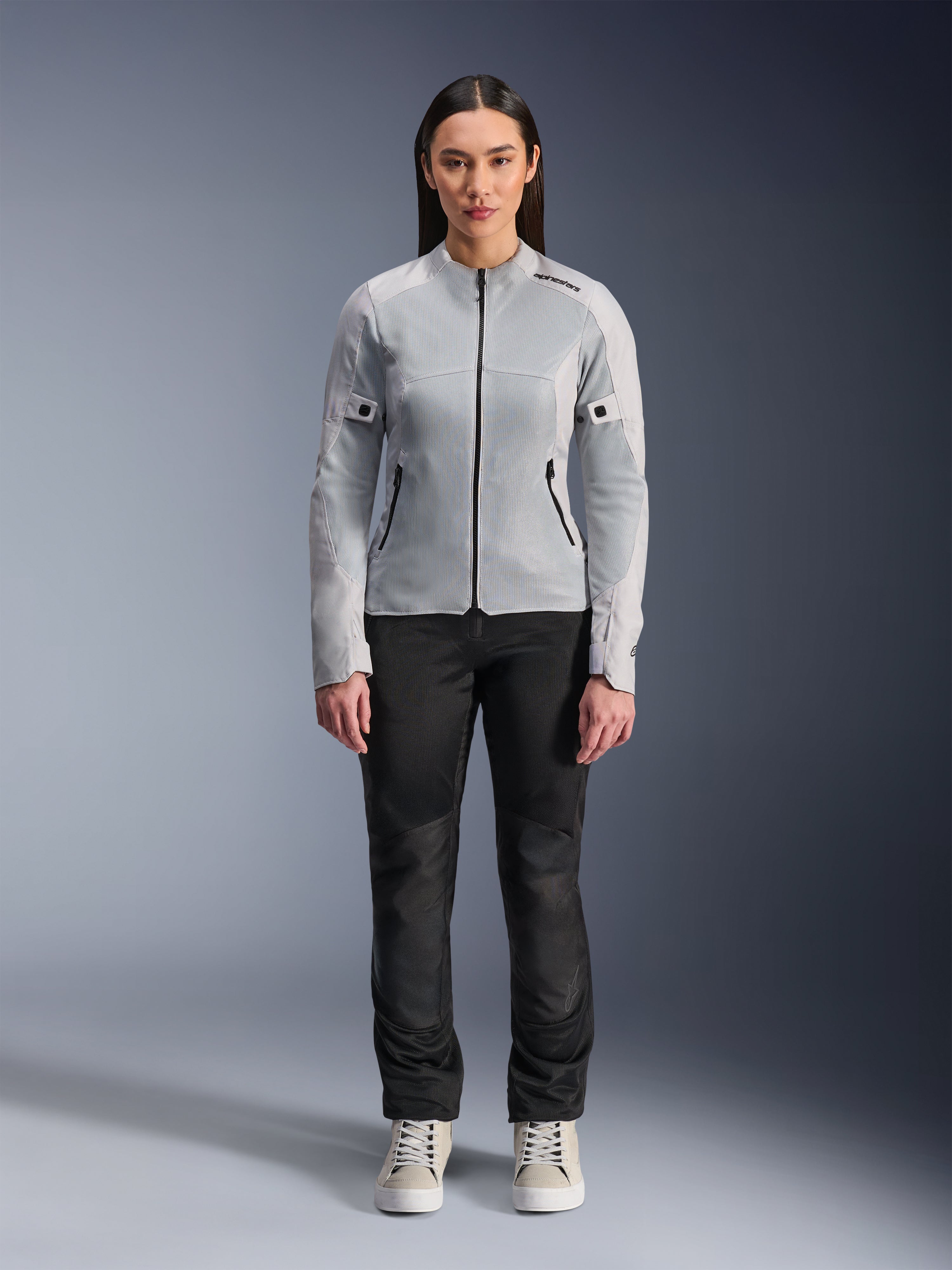 Frau in Alpinestars Stella C-1 Air Jacke in Silber, Vorderansicht, urbane Motorrad-Textiljacke mit Tech-Mesh-Einsätzen, kombiniert mit schwarzer Hose und weißen Sneakern, moderne Motorradausrüstung