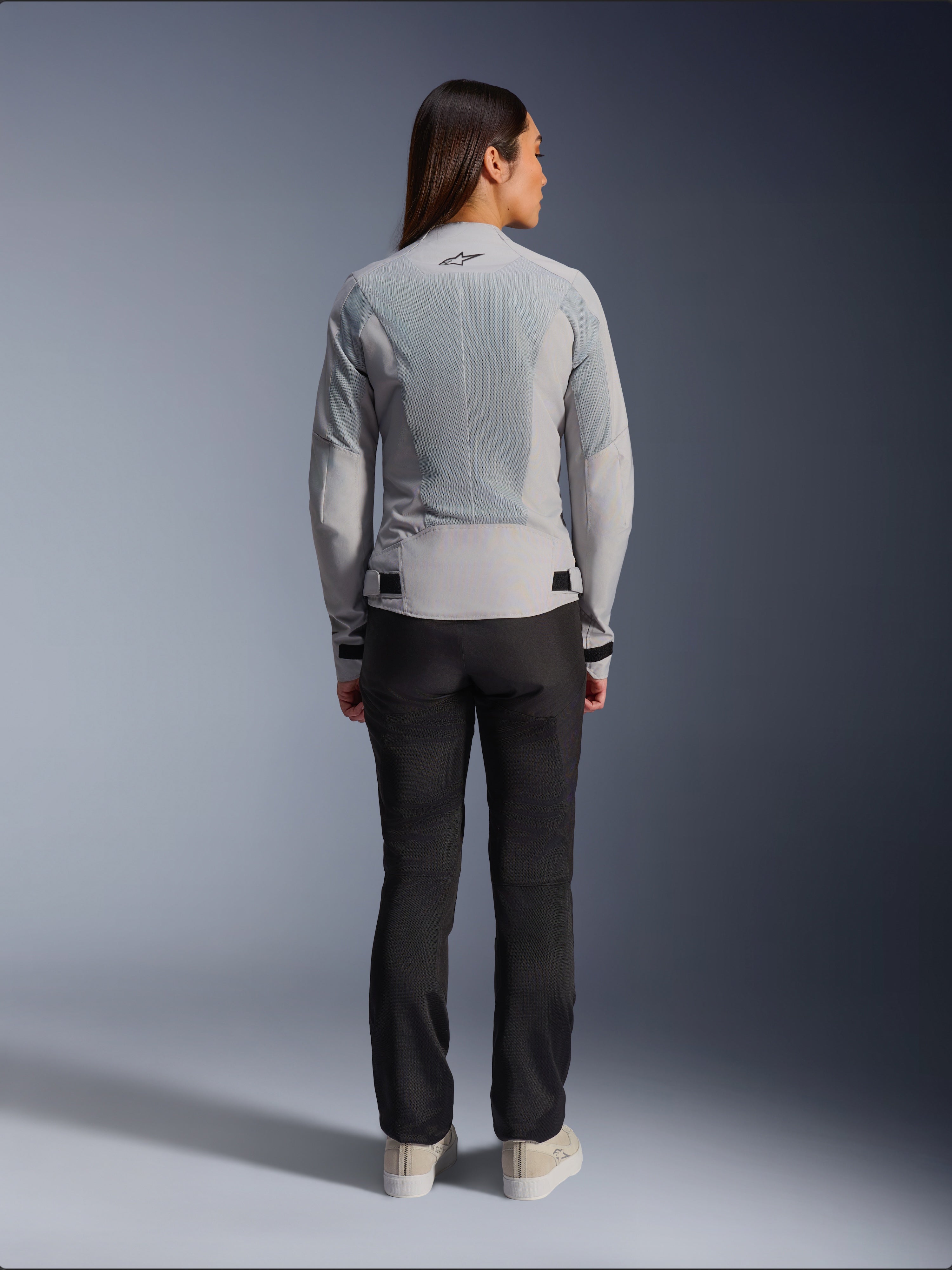 Frau in der Alpinestars Stella C-1 Air Jacke in Silber, Rückansicht, große Tech-Mesh-Einsätze zur Belüftung, reflektierendes Stern-Logo, kombiniert mit einer schwarzen Motorradhose und weißen Sneakern, moderne urbane Motorradausrüstung