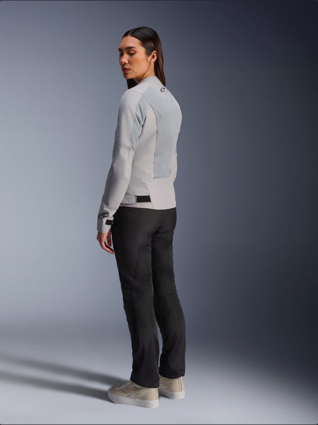 Frau trägt Alpinestars Stella C-1 Air Jacke in Silber, Rück- und Seitenansicht mit Tech-Mesh-Einsätzen und reflektierendem Schulter-Logo, kombiniert mit schwarzer Motorradhose und beigen Sneakern, moderne urbane Fahr-Ästhetik