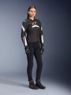 Weibliches Model trägt Alpinestars Stella T-GP Plus R V4 Jacke, Schwarz und Weiß, kombiniert mit schwarzer Motorradhose, Handschuhen und Stiefeln, schlankes Sport-Fit-Design für Motorradfahrerinnen
