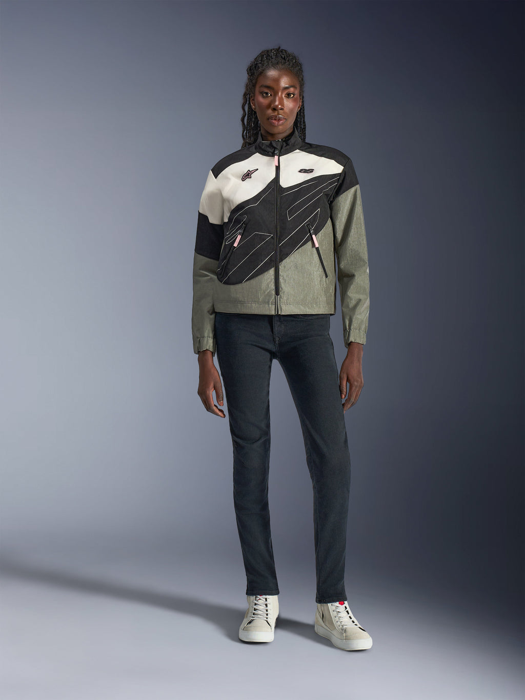 Alpinestars Stella Moshell Jacke, Damen-Motorradjacke, Schwarz, Pelzgrau und Off-White mit pinken Akzenten, getragen von einem Modell mit dunkler Jeans und cremeweißen Sneakern, asymmetrisches Reißverschluss-Design mit markanten geometrischen Grafiken, moderne urbane Motorradbekleidung