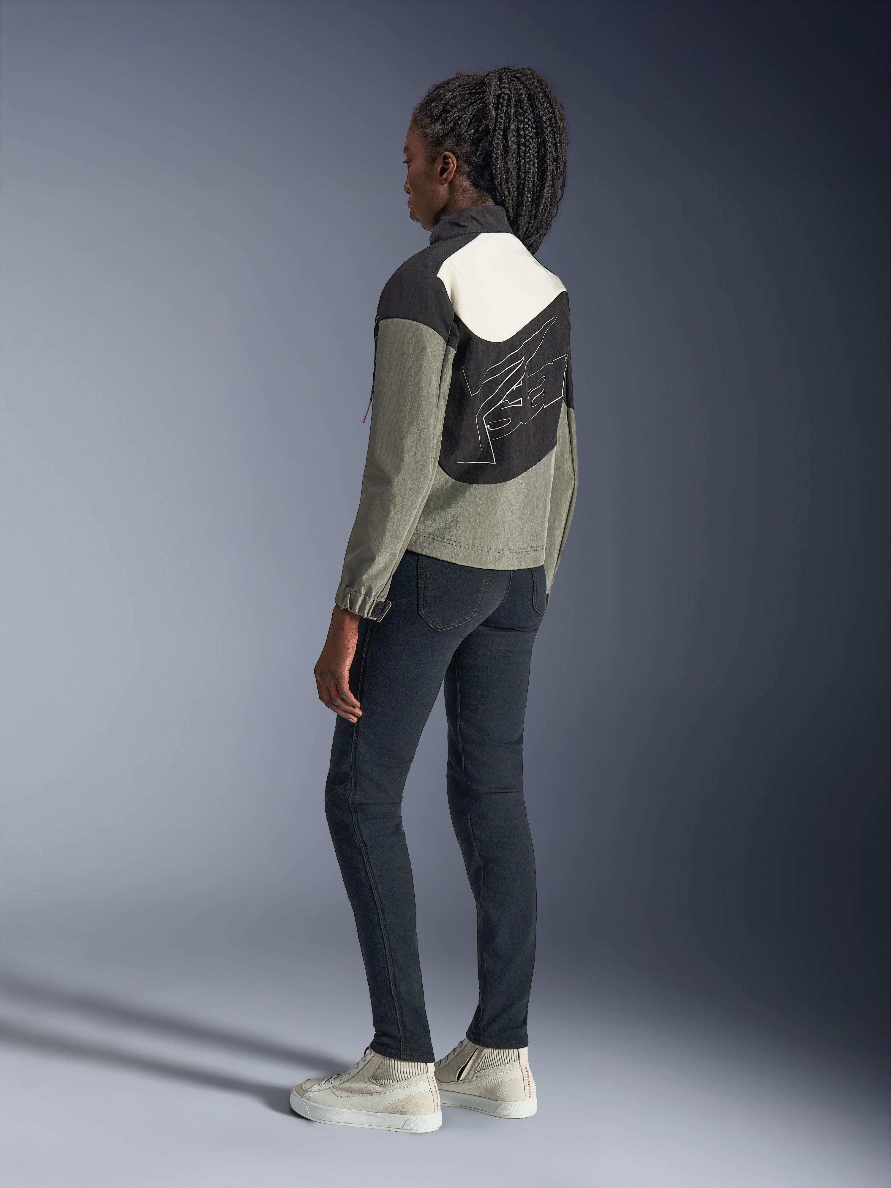 Alpinestars Stella Moshell Jacke für Damen, Urban-Motorradjacke, Farbkombination in Schwarz, Grau und Off-White, Rückansicht mit asymmetrischem Panel-Design und großer Grafik auf der Rückseite, kombiniert mit schwarzer Slim-Fit-Hose und Off-White Sneakern, moderne urbane Motorradausrüstung