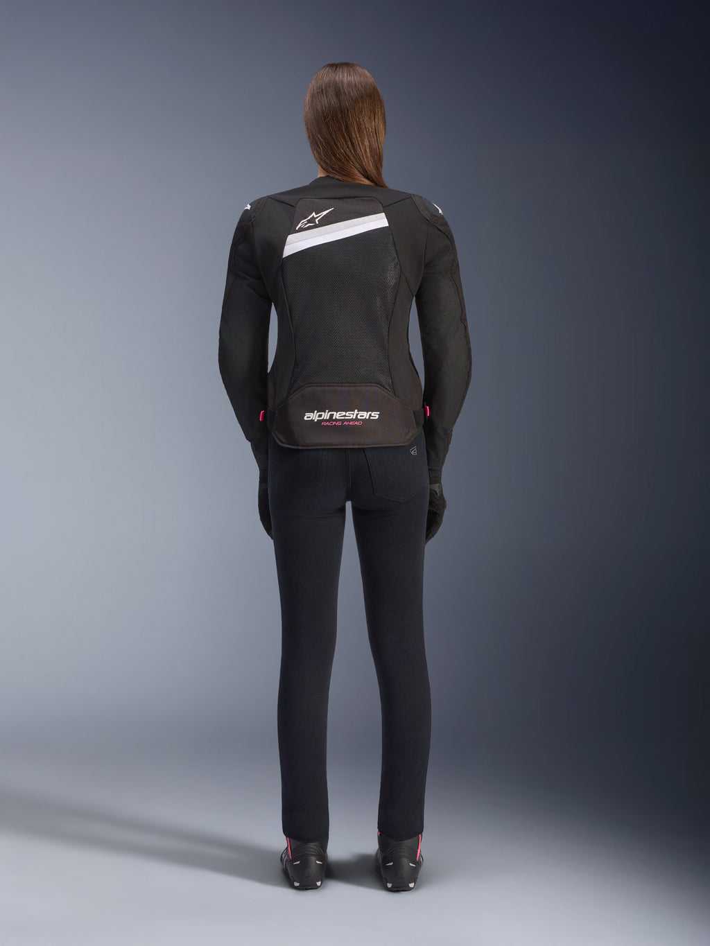 Alpinestars Stella T-GP Plus R V4 Airflow Jacket, Black and White, rear view showing extensive mesh ventilation panels and Alpinestars branding, worn by a female rider with slim-fit black motorcycle pants and boots, sport-fit textile gear for women