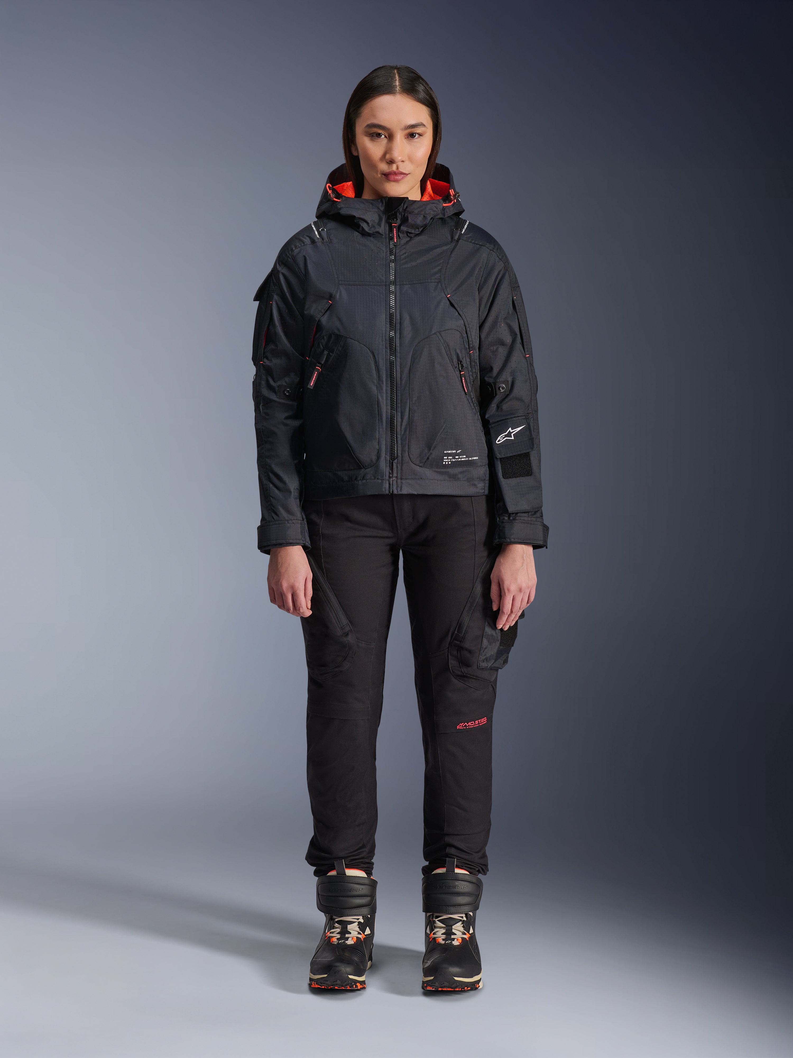 Weibliches Model in der Alpinestars Molly Air Jacke in Schwarz und Fluo Coral, Utility-inspiriertes Design mit Kapuze und korallenfarbenem Innenfutter, kombiniert mit schwarzer Motorradhose und Stiefeln, urbane zeitgenössische Motorradausrüstung