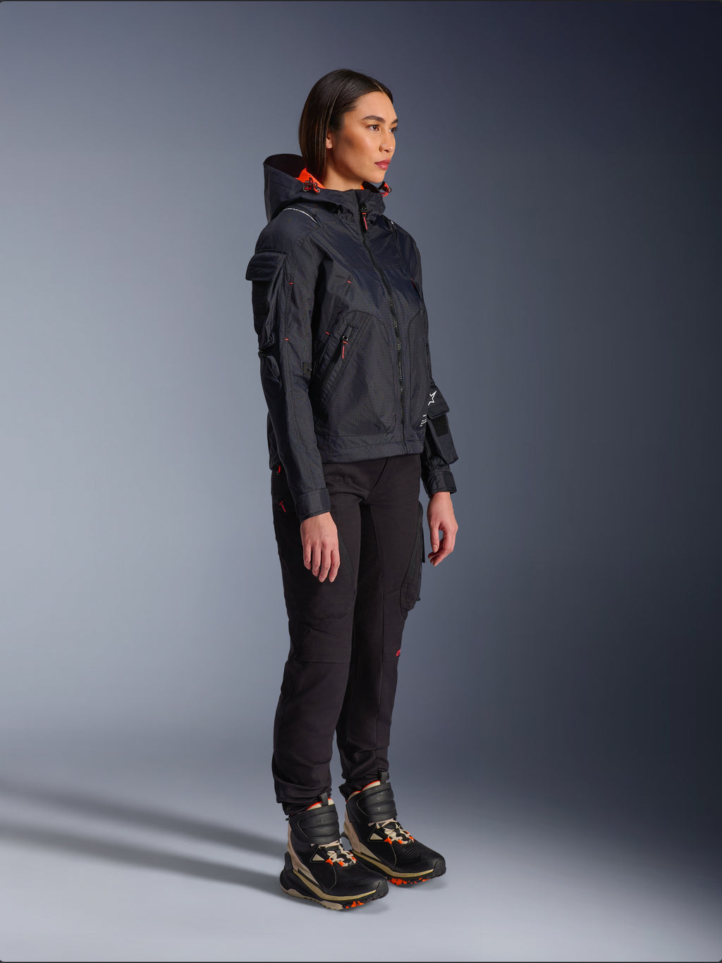 Motorradfahrerin in der Alpinestars Molly Air Jacke in Schwarz Fluo Coral, leichte Textil-Motorradjacke mit leuchtend orangefarbenem Kapuzenfutter, kombiniert mit schwarzer Motorradhose und technischen Stiefeln, urbanes zeitgenössisches Utility-inspiriertes Design.
