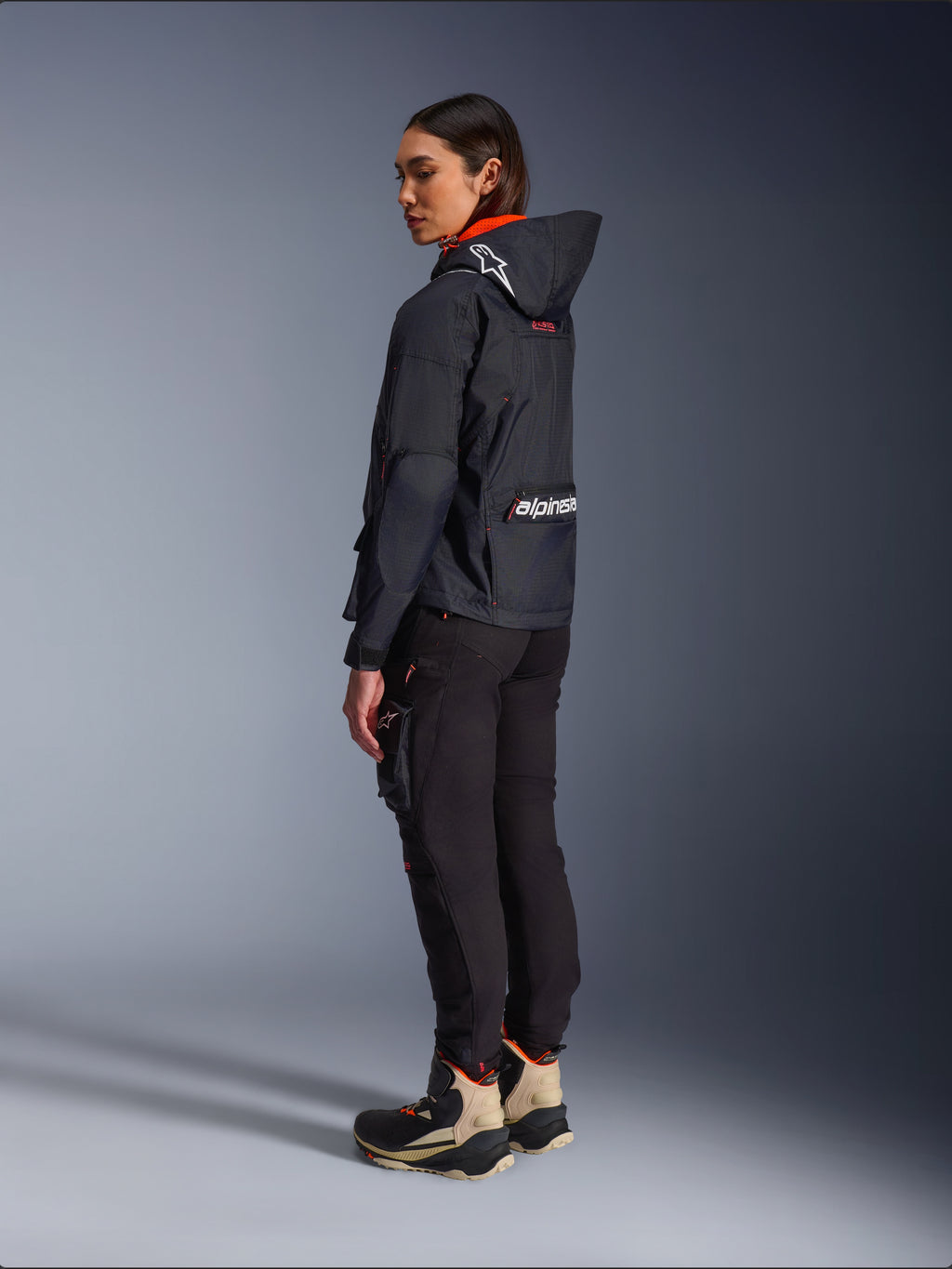 Alpinestars Actea Damen-Hose, Schwarz, moderne Motorradhose mit schmal zulaufender Passform und Cargo-Tasche am Oberschenkel, Seitenansicht von hinten an einem Modell mit dunkler Kapuzenjacke und Motorradstiefeln, urbaner Fahrstil.