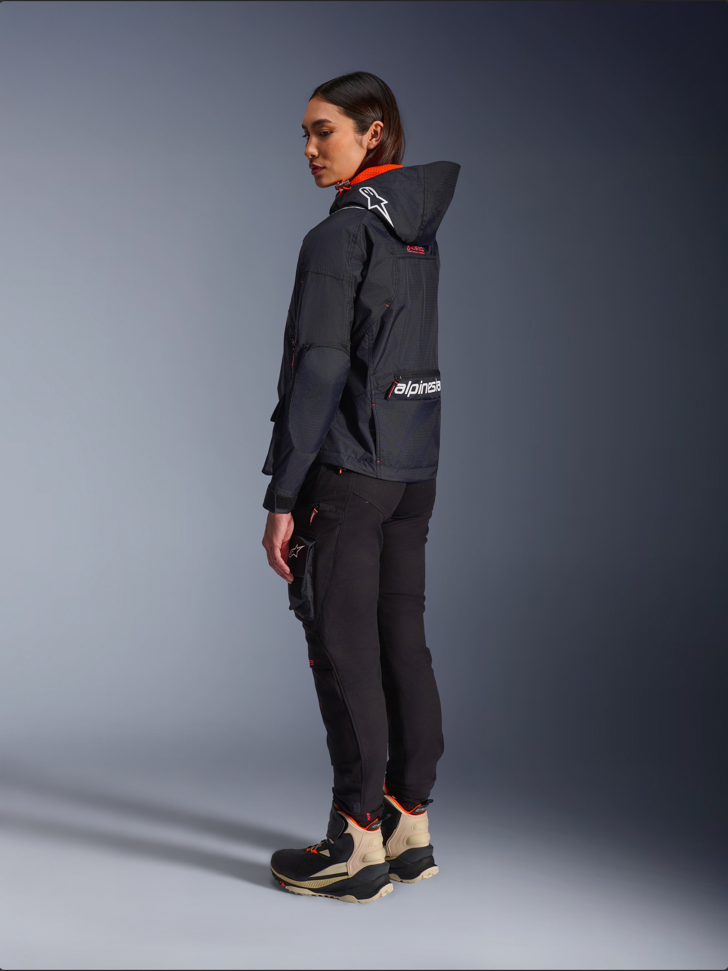 Alpinestars Actea Damen-Hose, Schwarz, moderne Motorradhose mit schmal zulaufender Passform und Cargo-Tasche am Oberschenkel, Seitenansicht von hinten an einem Modell mit dunkler Kapuzenjacke und Motorradstiefeln, urbaner Fahrstil.