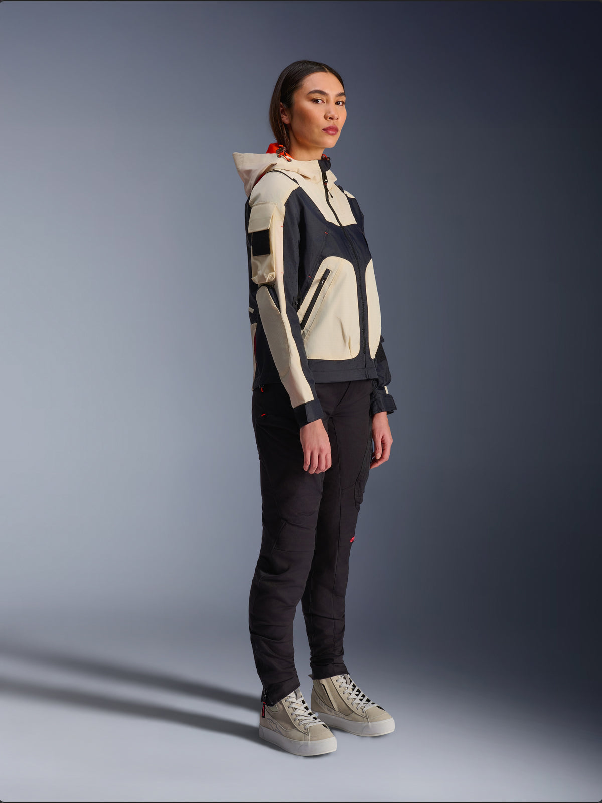 Alpinestars Molly Air Jacke in Black Almond, getragen von einer Fahrerin mit schwarzer Hose und cremefarbenen High-Top-Sneakern, zweifarbiges Design mit schwarzen und beigen Panels, integrierte Kapuze, moderner urbaner Stil.