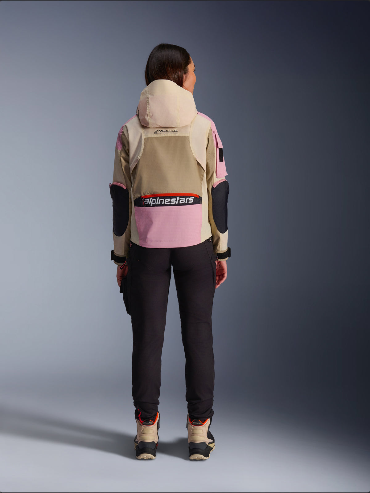 Motorradfahrerin in der Rückansicht, trägt die Alpinestars Molly Air Jacke in Sand Pink Almond, mit beiger Kapuze und rosa Einsätzen mit schwarzen Akzenten, kombiniert mit einer schwarzen Motorradhose und beigen Motorradstiefeln, urbane zeitgemäße Motorradbekleidung